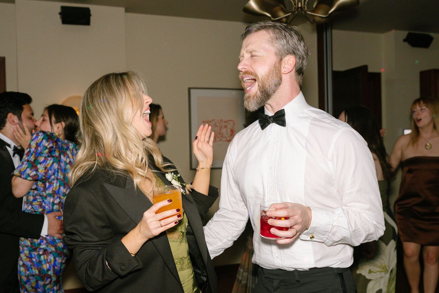Two party guests share a laugh while holding drinks at a social gathering.
