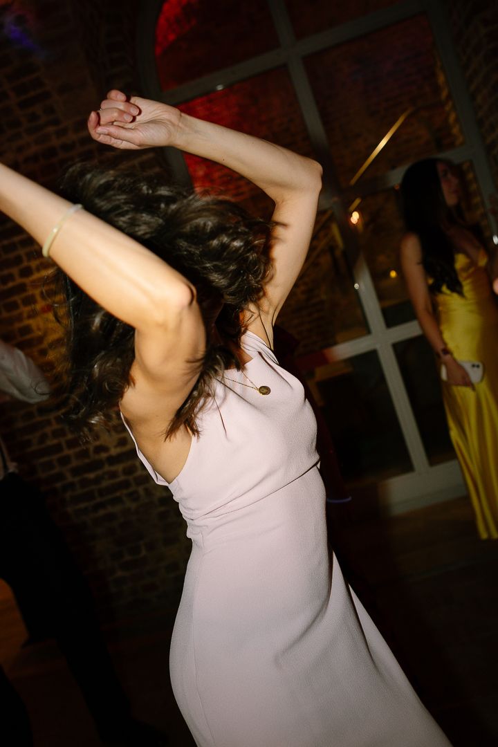Person in white tank top dancing energetically in dimly lit room with brick walls and ambient lighting.