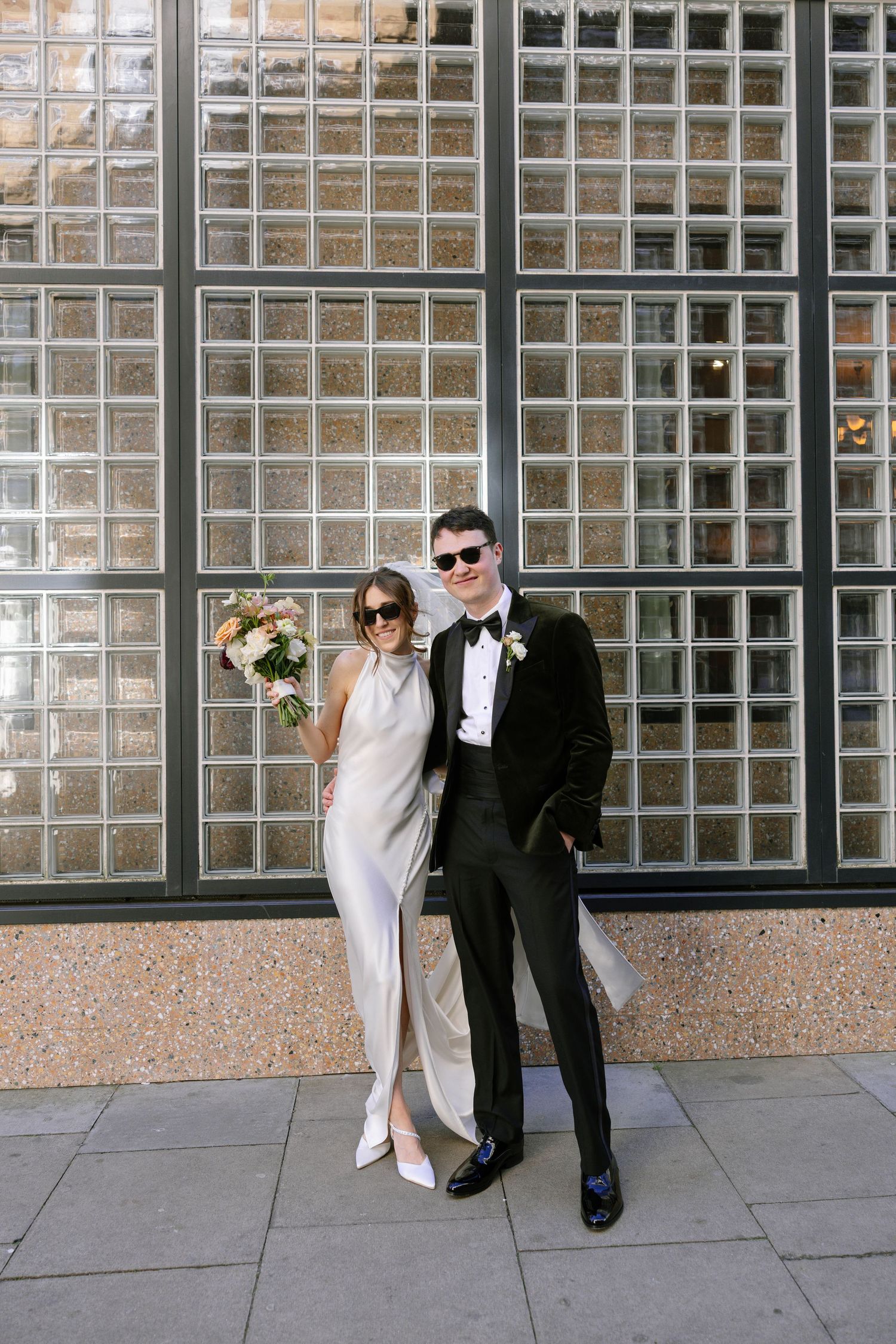 Wedding couple posing in formal attire against a glass block wall.