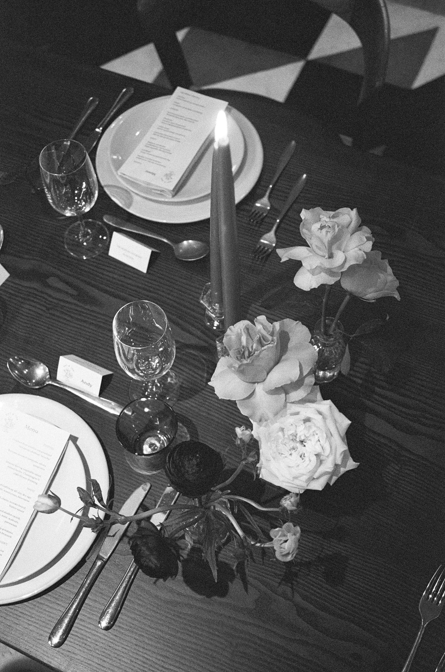 Black and white photograph of an elegant table setting with wine glasses, place settings, and roses in a vase.