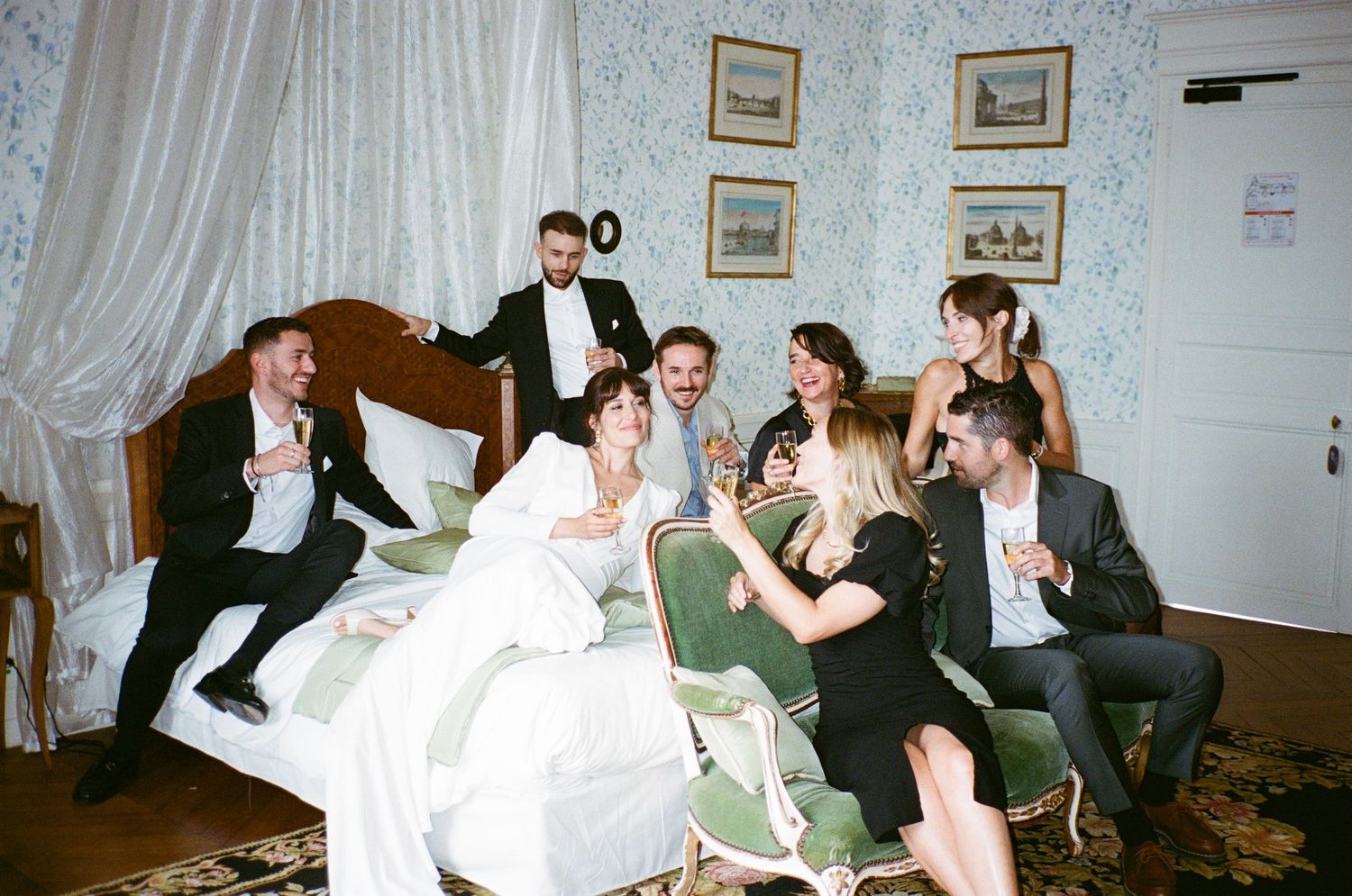 A group of formally dressed people gather in a bedroom, sharing laughter and conversation at what appears to be a wedding event.