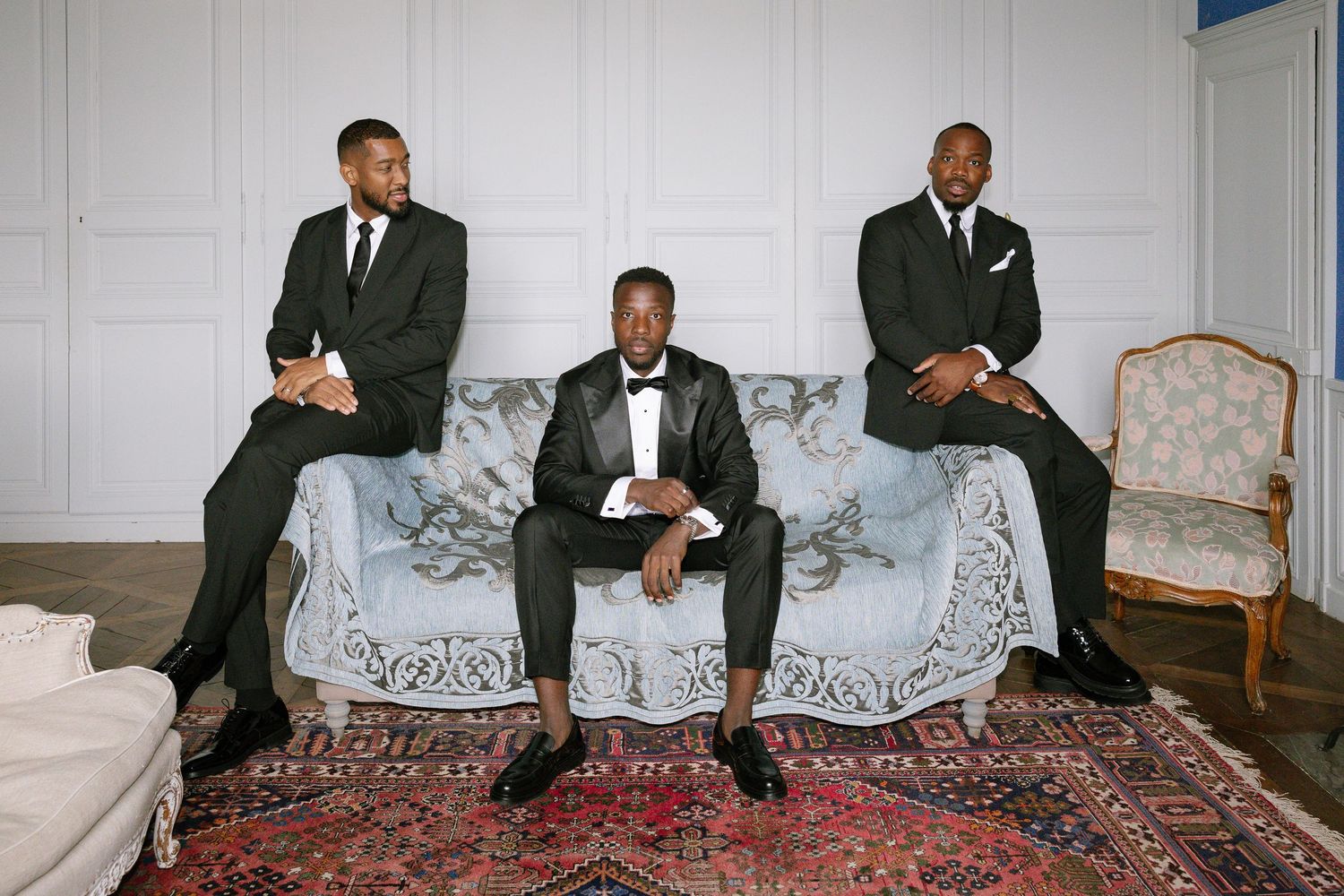 Five formally dressed men pose at various positions on a patterned floor rug in a light colored room.