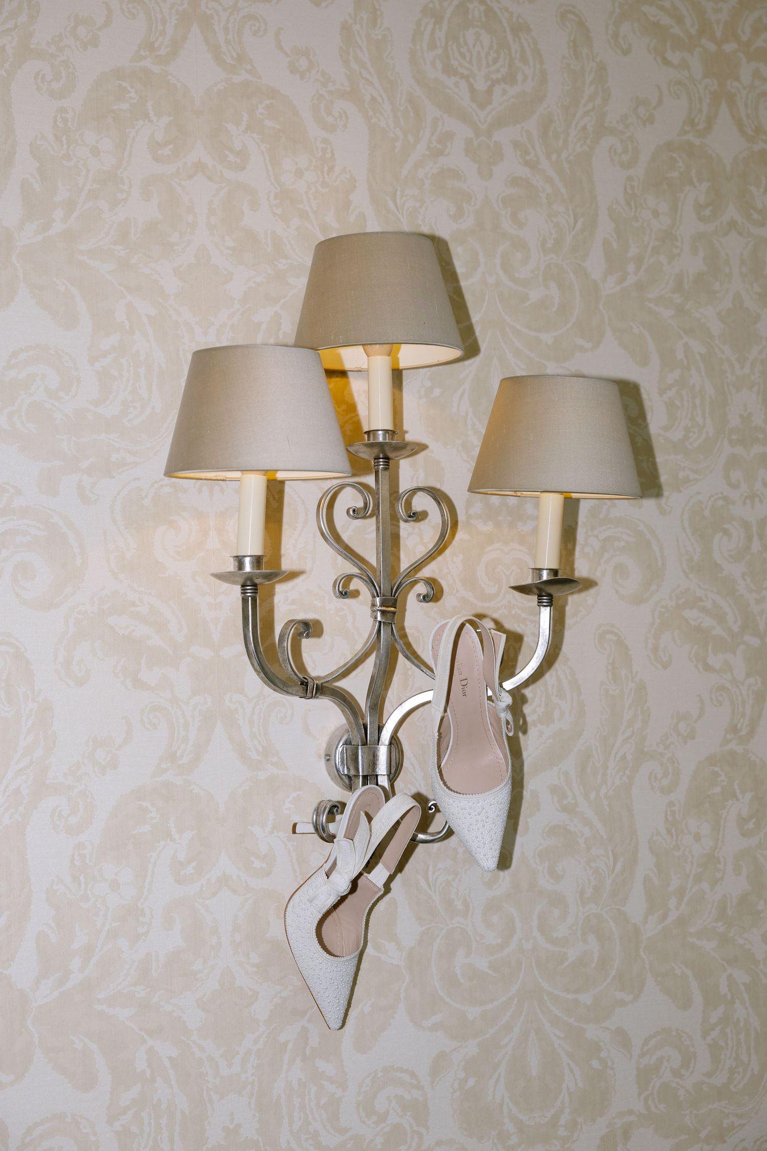 Elegant wall sconce with three lampshades and decorative metalwork against damask wallpaper.