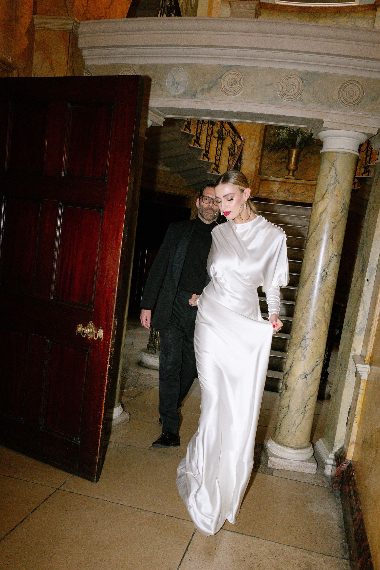 Elegant couple in formal attire walking through ornate hallway with marble columns.