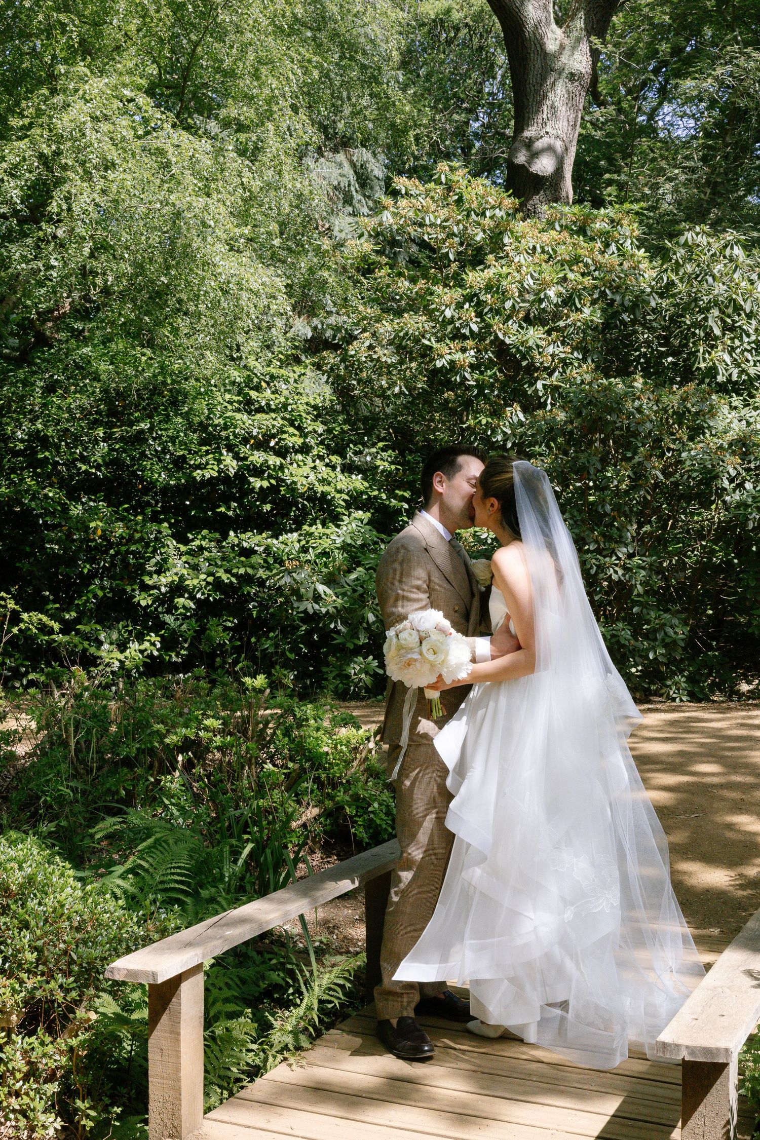 Bride and groom share a kiss on a wooden bridge surrounded by lush green trees on their wedding day.