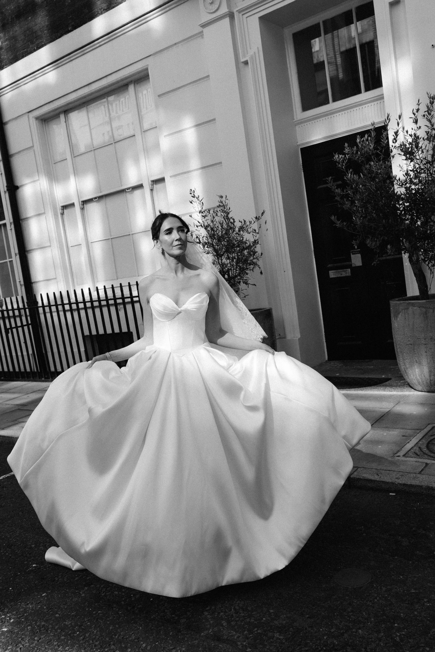 A bride in a stunning strapless ballgown with voluminous skirt sits elegantly on steps outside a white building.