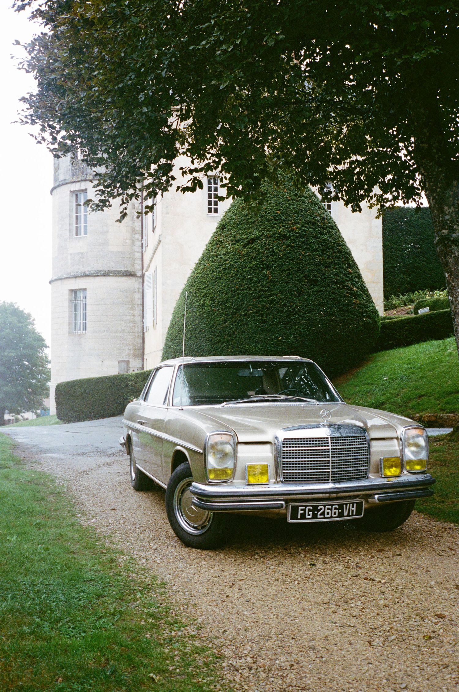 A vintage silver Mercedes-Benz sedan parked on a gravel driveway near a French chateau with manicured hedges.