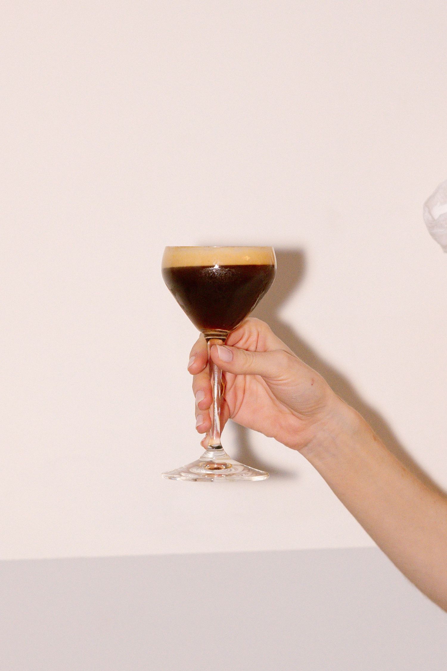 Hand holding an espresso martini cocktail against pale pink background.