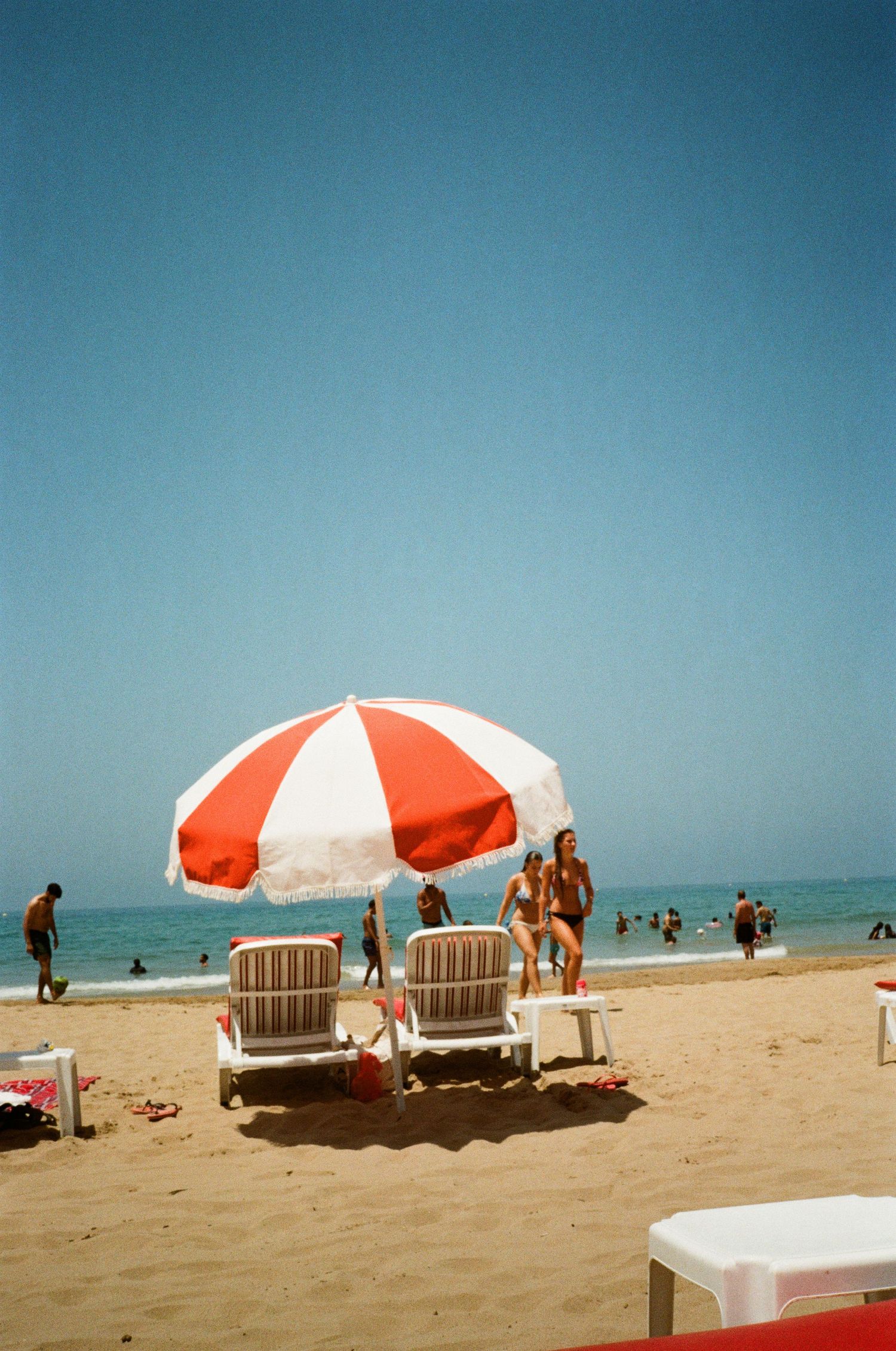 Red and white striped beach umbrella shades two lounge chairs on a sandy beach with blue sky and ocean in background in the French Riviera.