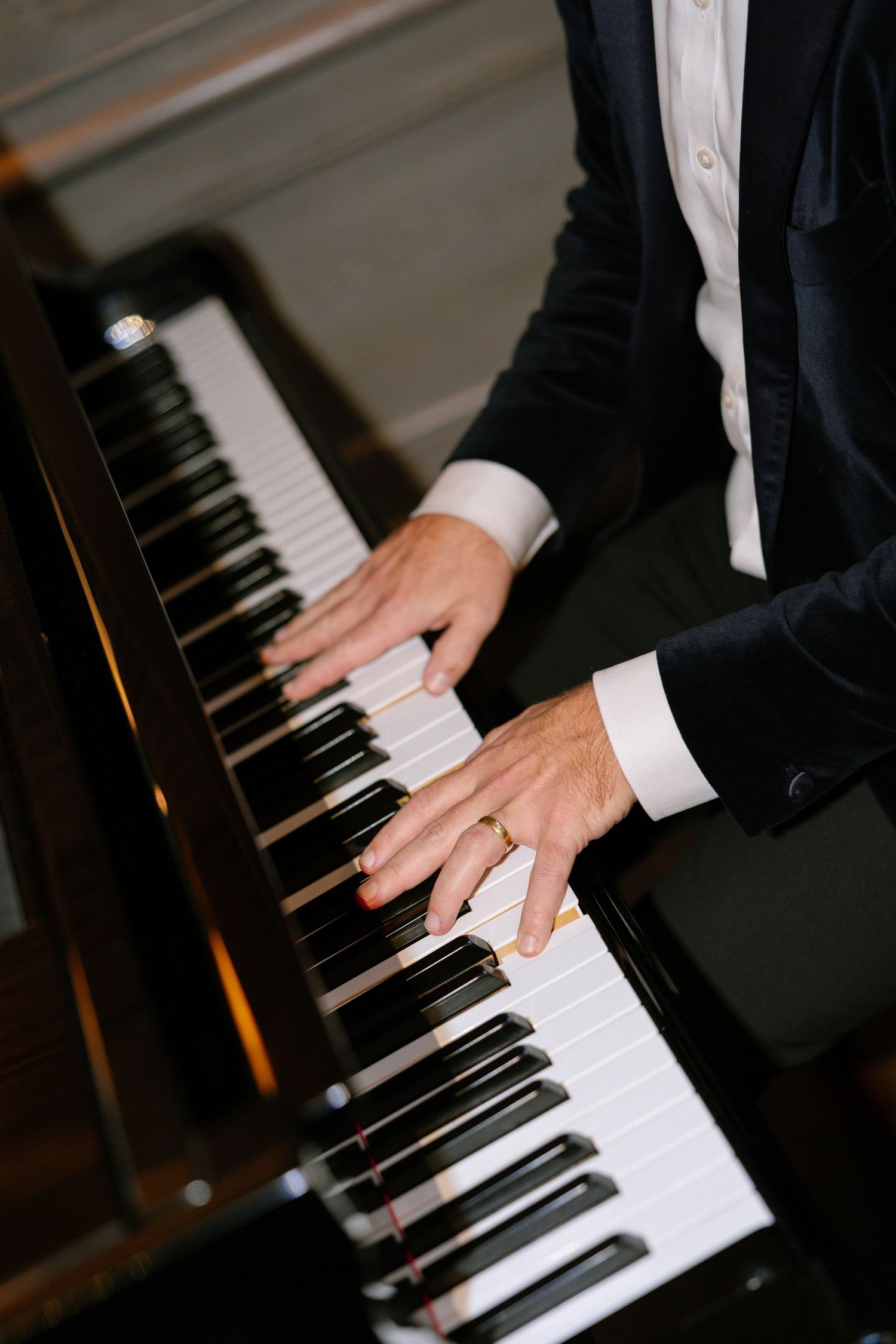 Close-up of hands playing piano keys while wearing formal attire with wedding ring visible.