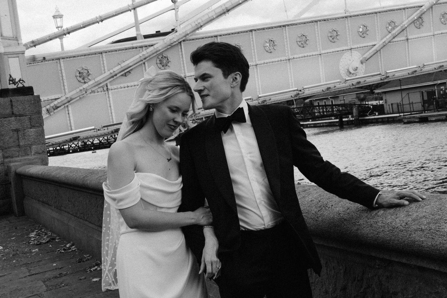 A romantic black and white photo of a couple in formal attire standing by a bridge in an urban setting.