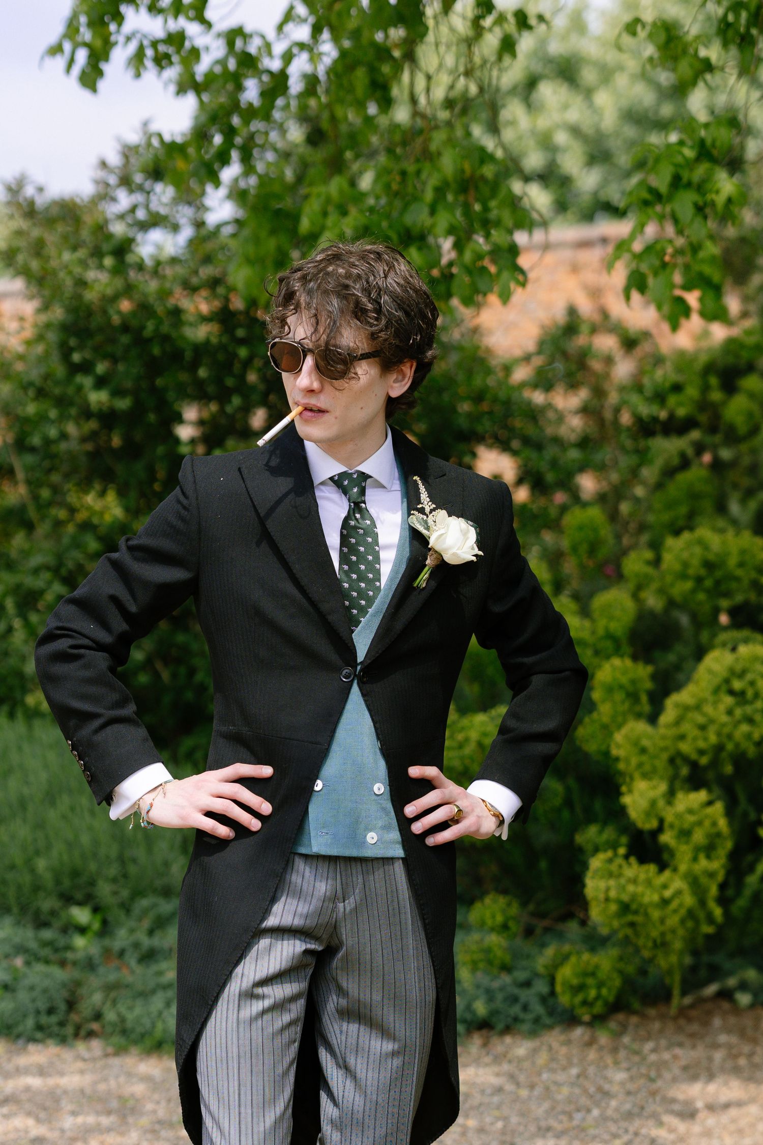A formal morning suit with tails and pinstripe trousers worn with sunglasses and a boutonniere outdoors.