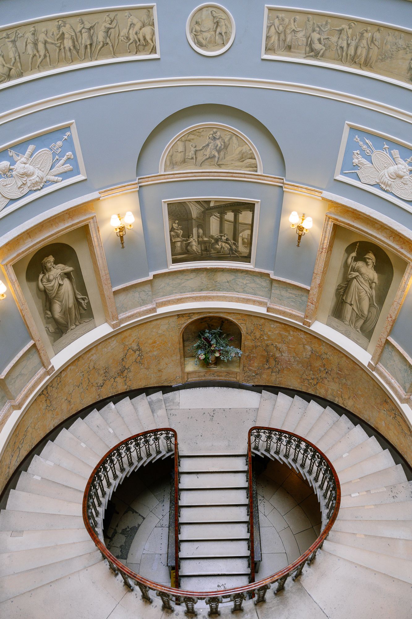Elegant curved staircase with ornate neoclassical artwork and reliefs adorning the domed ceiling in a historic building.