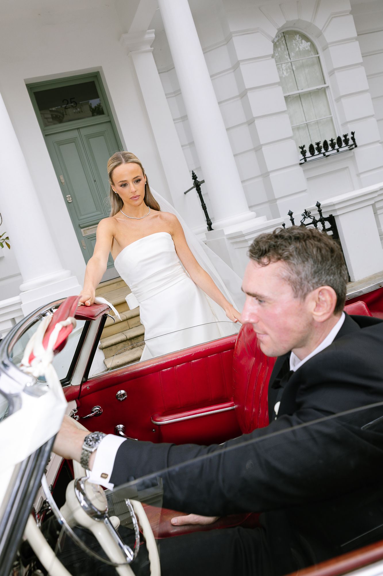 A classic red vintage car sits in front of a grand white building with columns and an elegant staircase during a wedding.