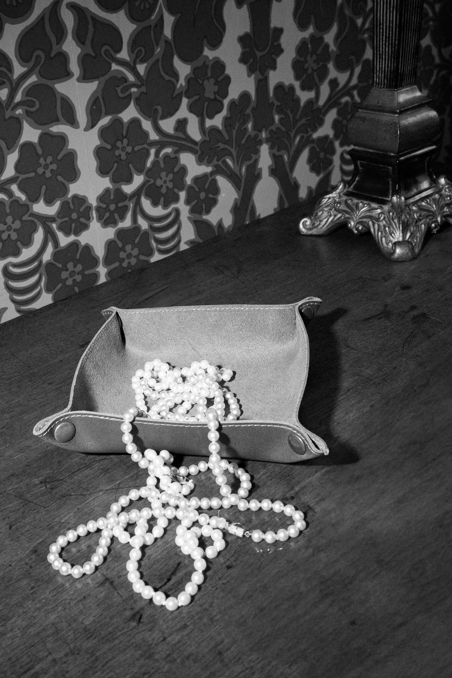 Black and white photo of pearl necklace in leather valet tray against floral wallpaper.