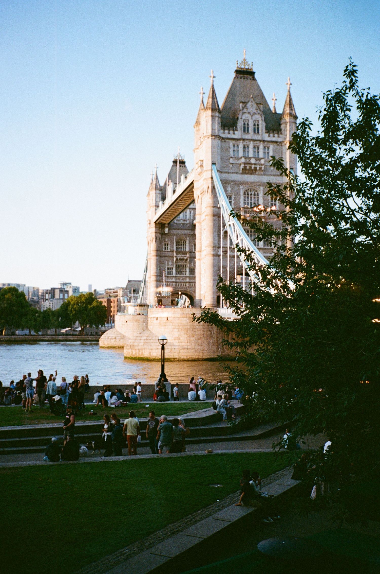 Tourists gather near London's iconic Tower Bridge at dusk as the historic landmark illuminates against the evening sky.