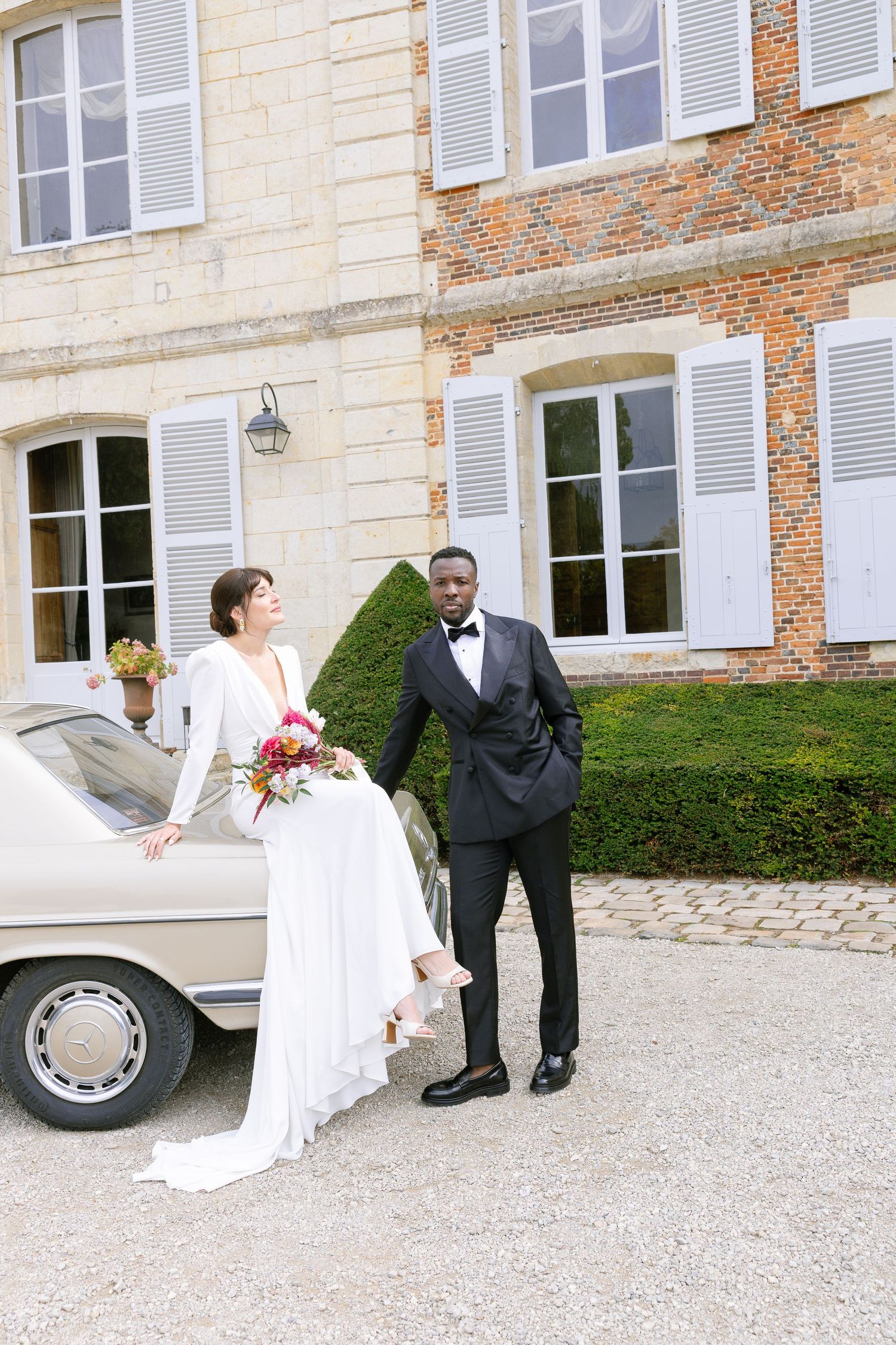 A bride and groom stand next to a vintage car outside an elegant French chateau with white shuttered windows.