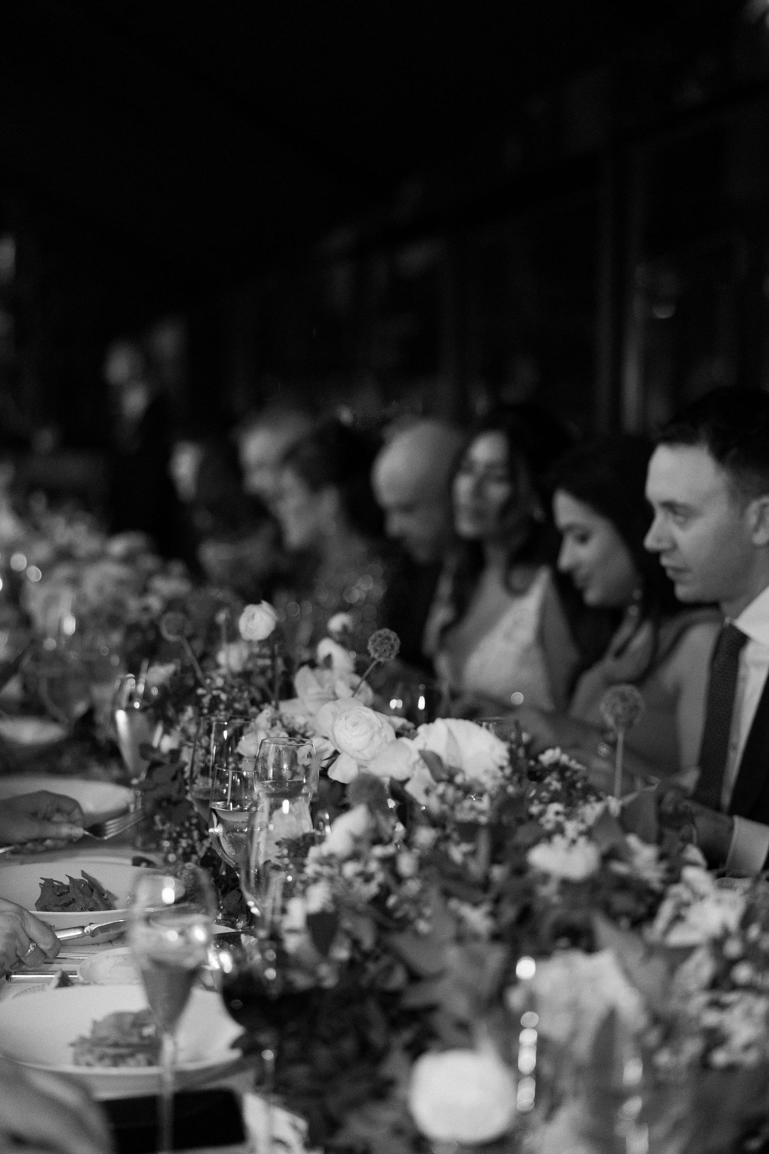 Black and white photograph of guests seated at an elegant wedding reception table with floral centerpieces.