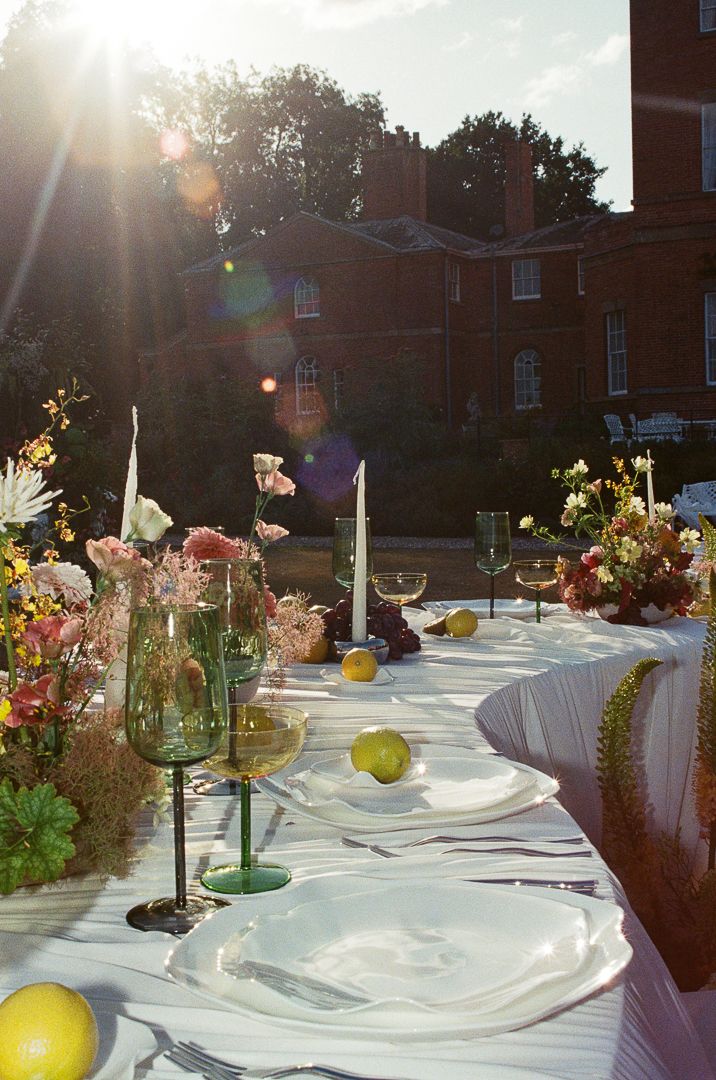 An elegant outdoor table setting with white linens, candles, and wildflowers backlit by golden sunlight at dusk.