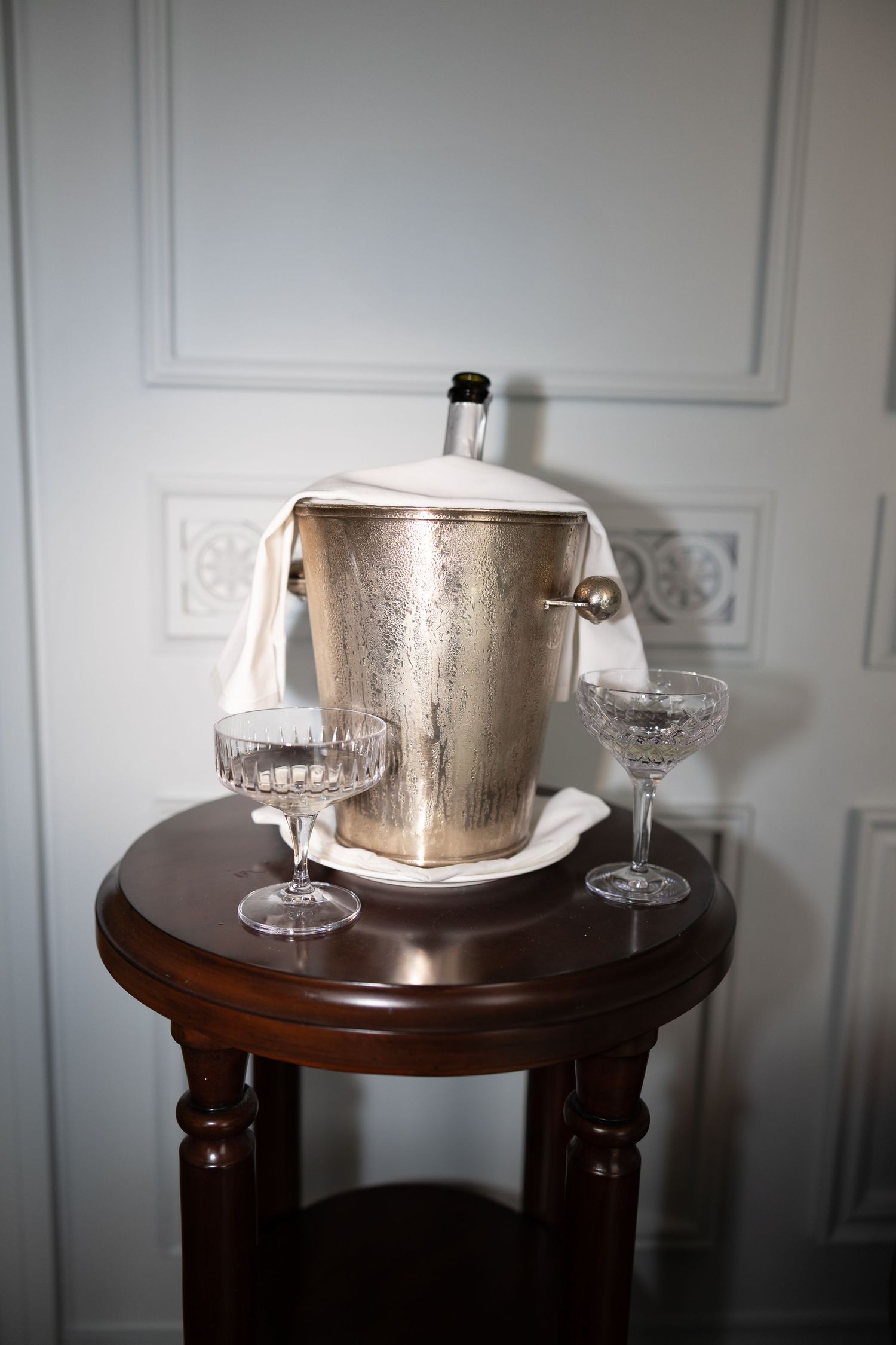 Silver champagne bucket and crystal glasses sit elegantly on a dark wooden side table against a white wall.