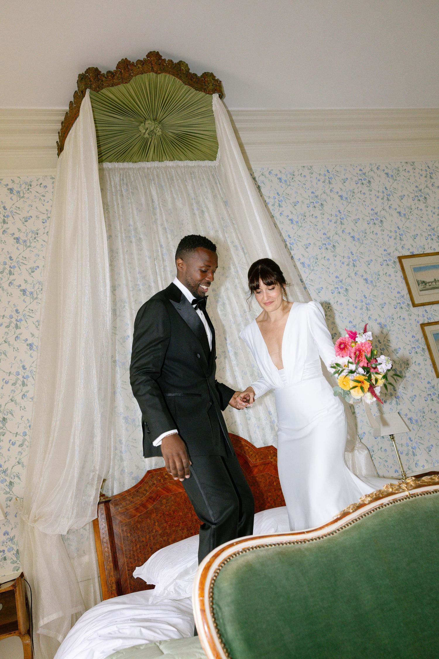 A couple in white dress and black suit share an intimate moment on a green antique sofa in a vintage-style room.