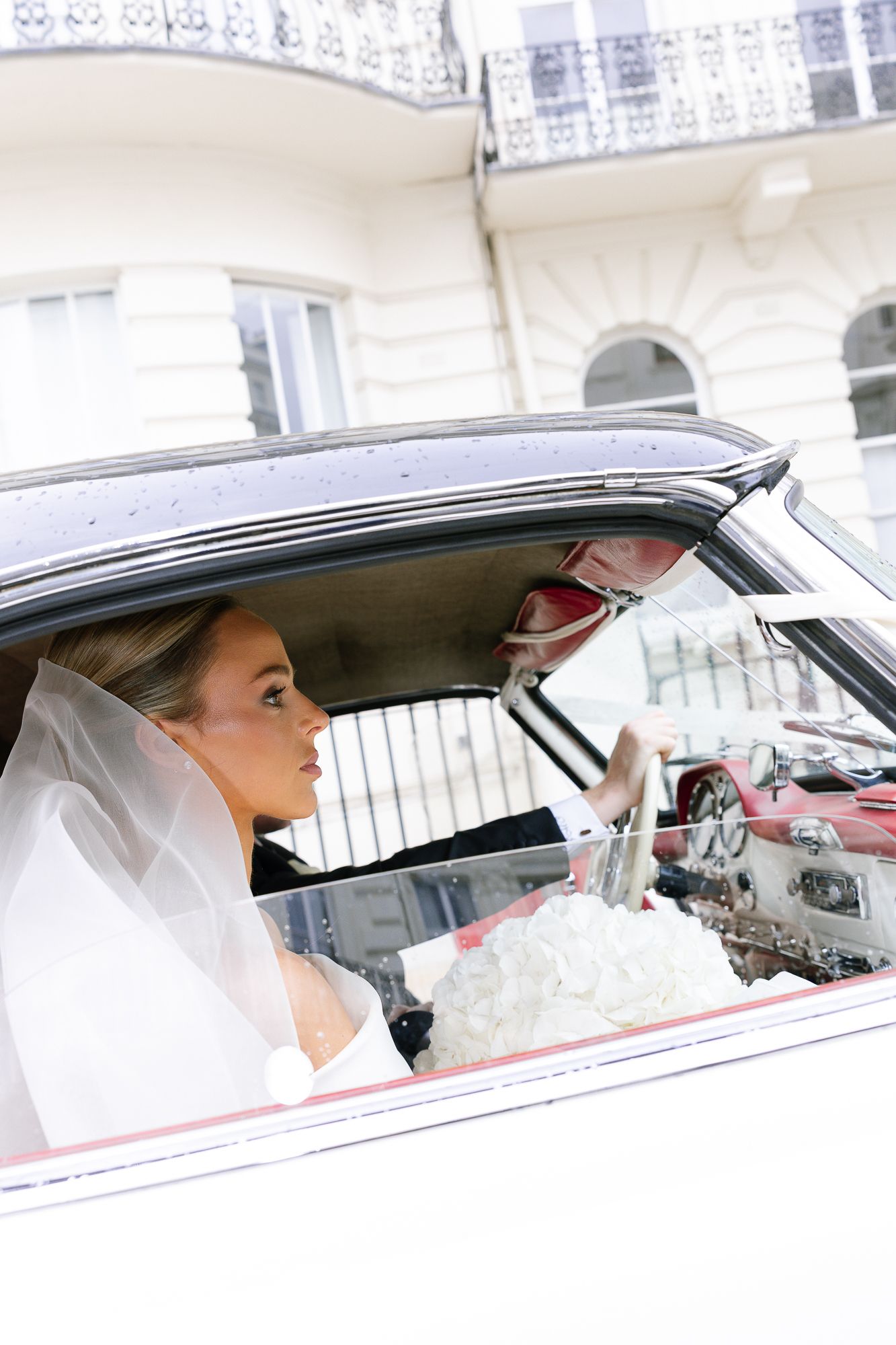 A bride wearing a veil sits in a vintage white car looking out the window on her wedding day.
