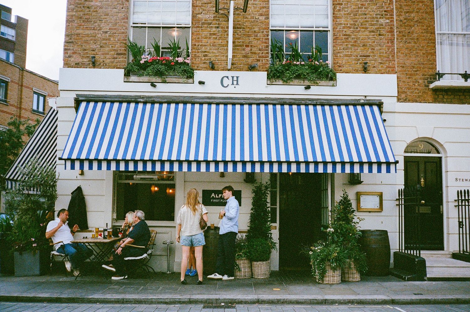 A row of charming brick storefronts with blue and white striped awnings line a street with pedestrians walking by.