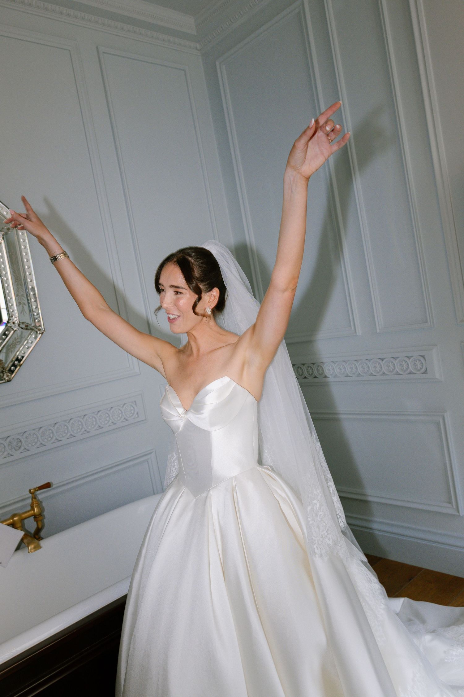 A graceful figure in a flowing white wedding dress dances joyfully with raised arms in an elegant room.