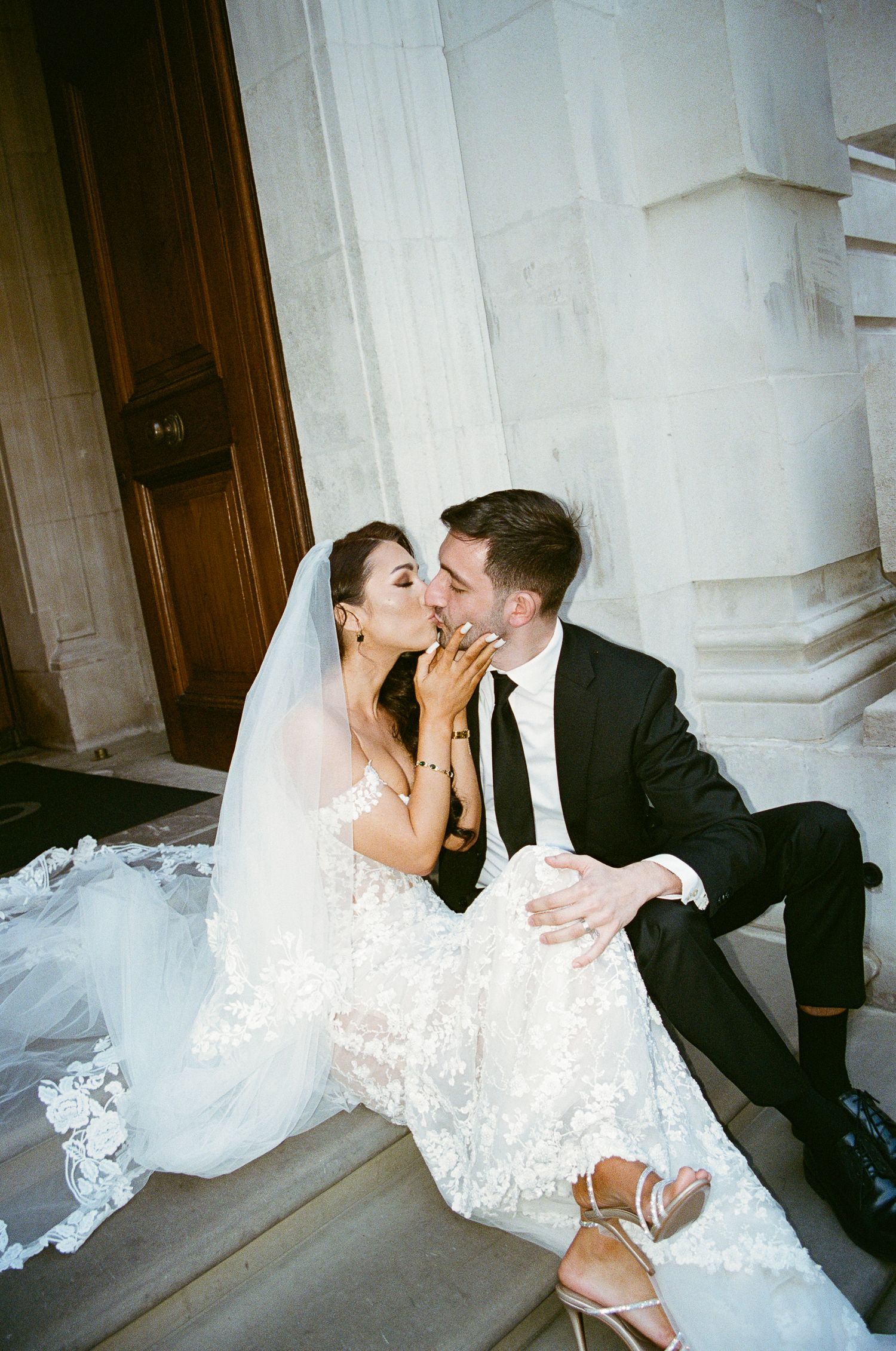A bride and groom share a romantic kiss on stone steps, her lace gown and veil flowing beside them.