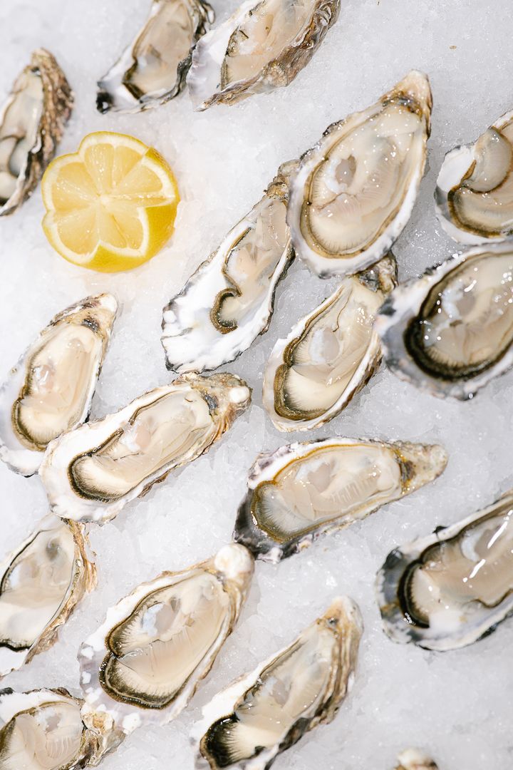 Fresh shucked oysters on ice arranged in diagonal rows with lemon wedges on a light colored surface.