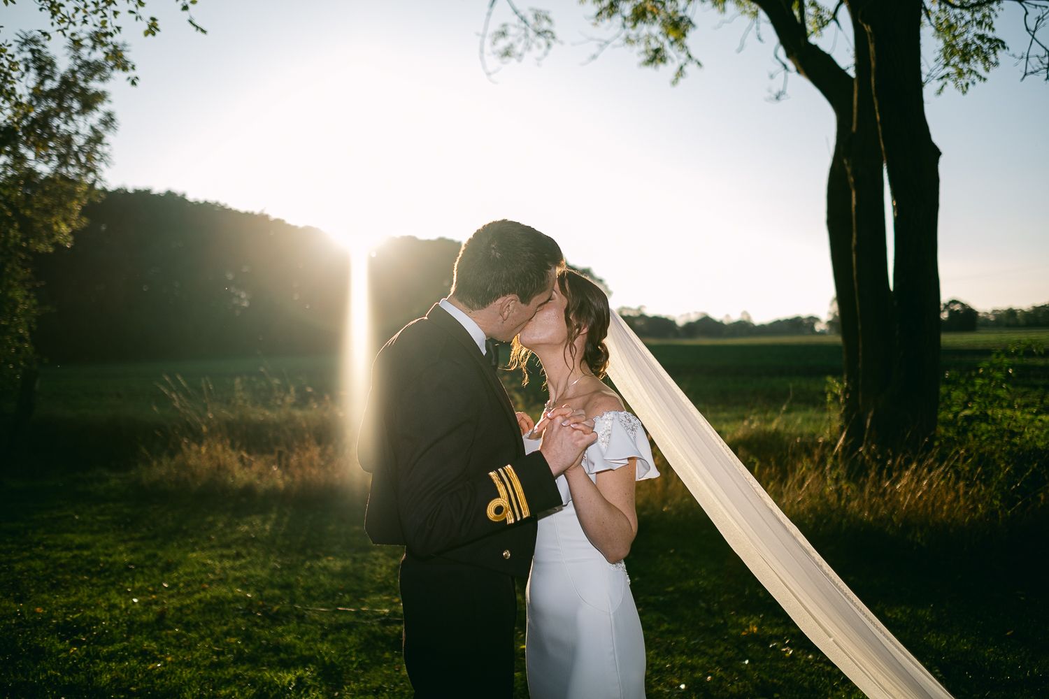 A couple embraces in a sunlit field at golden hour as a wedding veil floats in the breeze.