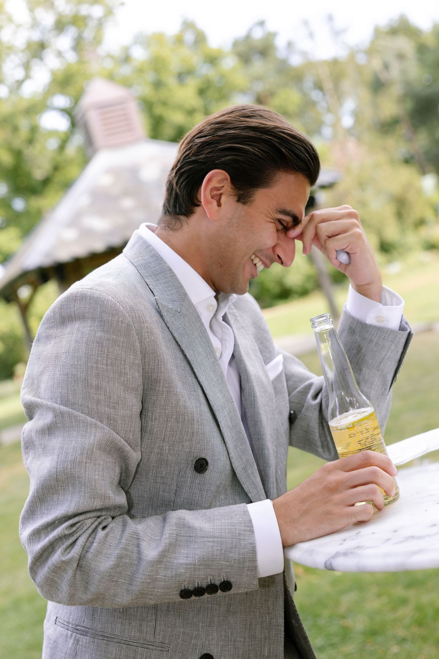 Man in grey suit laughing while holding a drink outdoors.