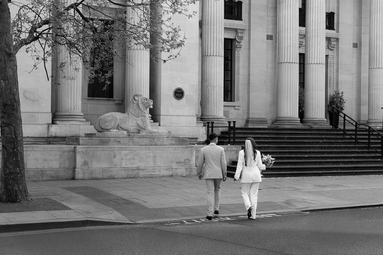 Two people walk past a grand building with columns and stone steps in a classic black and white photograph.