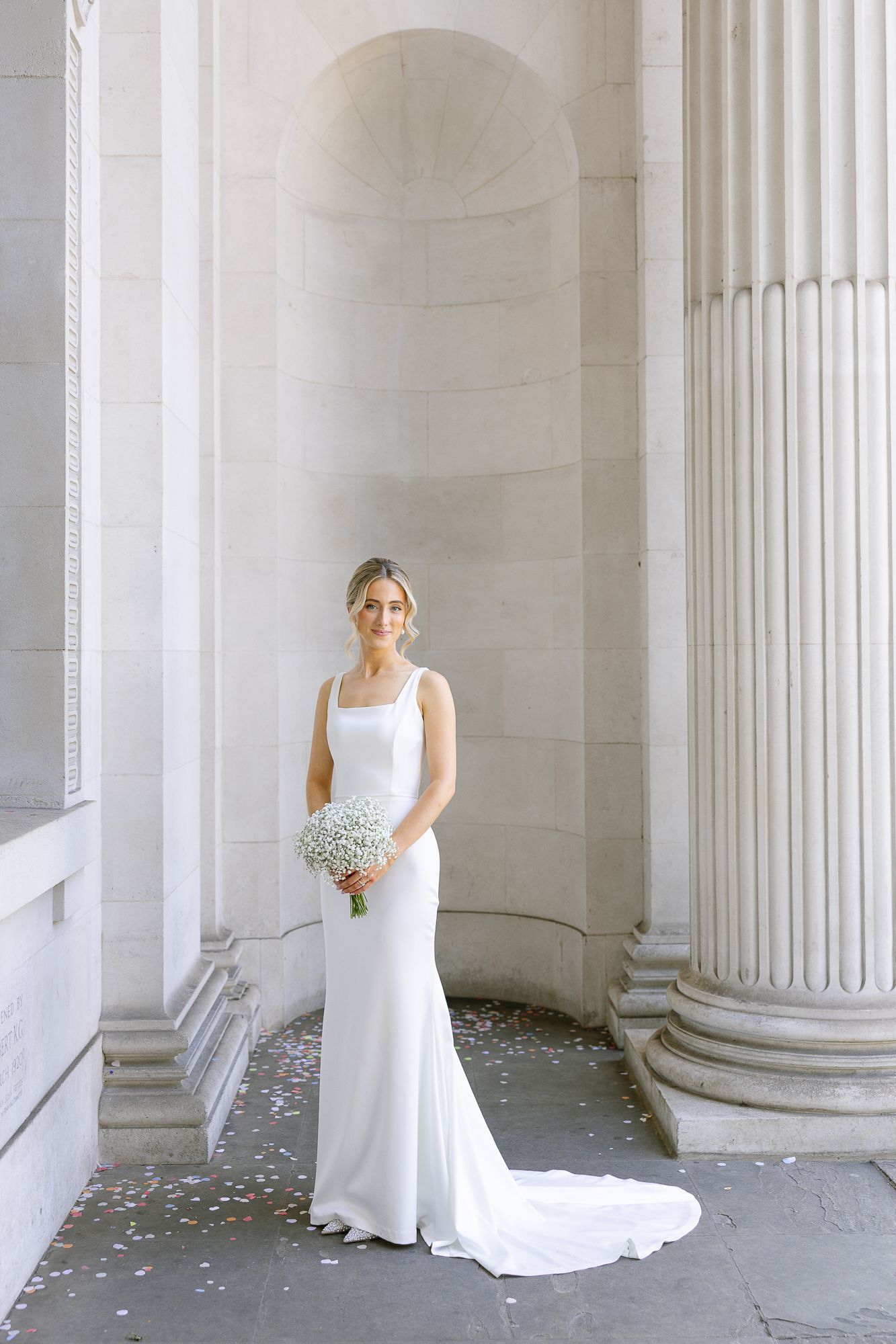 Elegant bride in white dress stands between classical white columns with flower petals scattered on the ground.