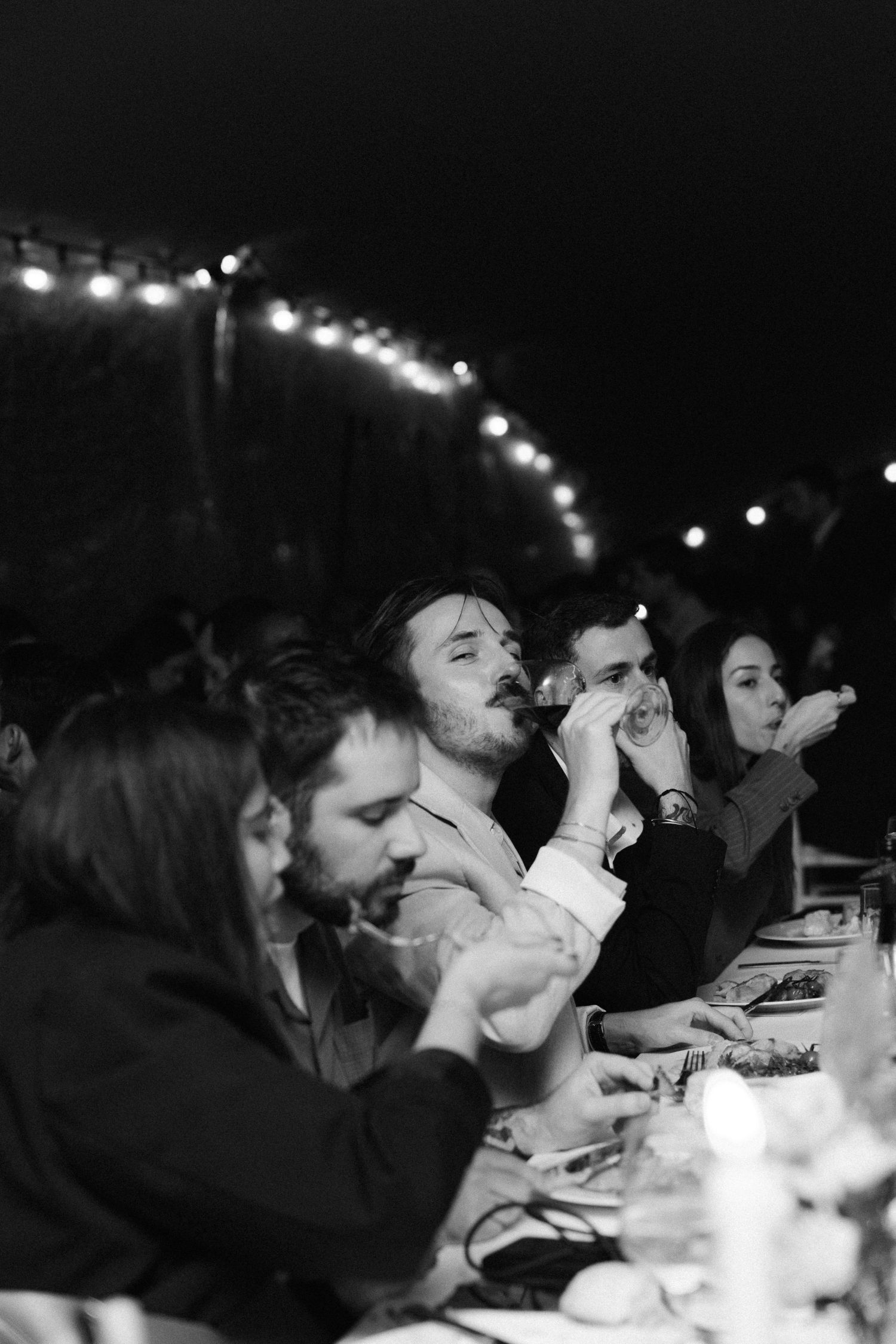 A black and white photo shows people drinking beverages at a dimly lit gathering with string lights overhead. Wedding Reception.