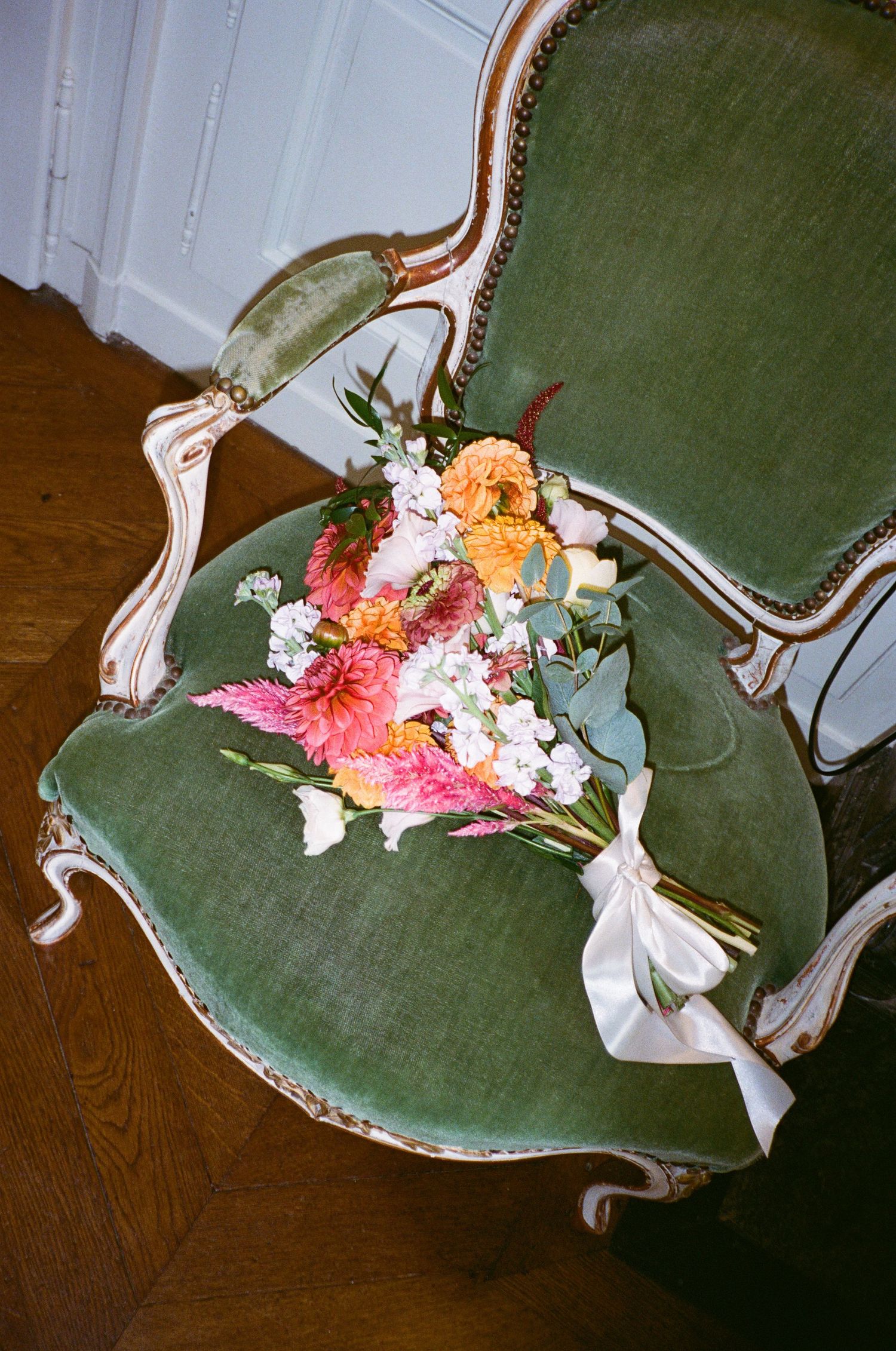A vintage green velvet chair decorated with colourful floral bridal bouquet arrangements and white ribbon against a wooden floor.