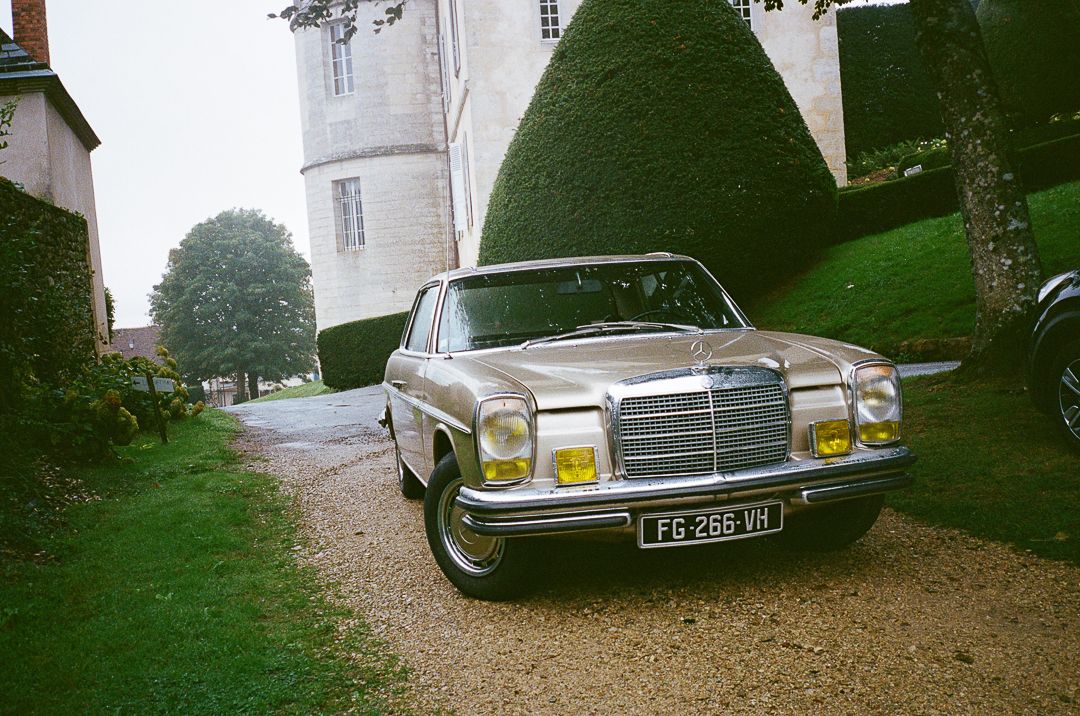 A classic Mercedes-Benz sedan parked in front of a white building with manicured topiary bushes.