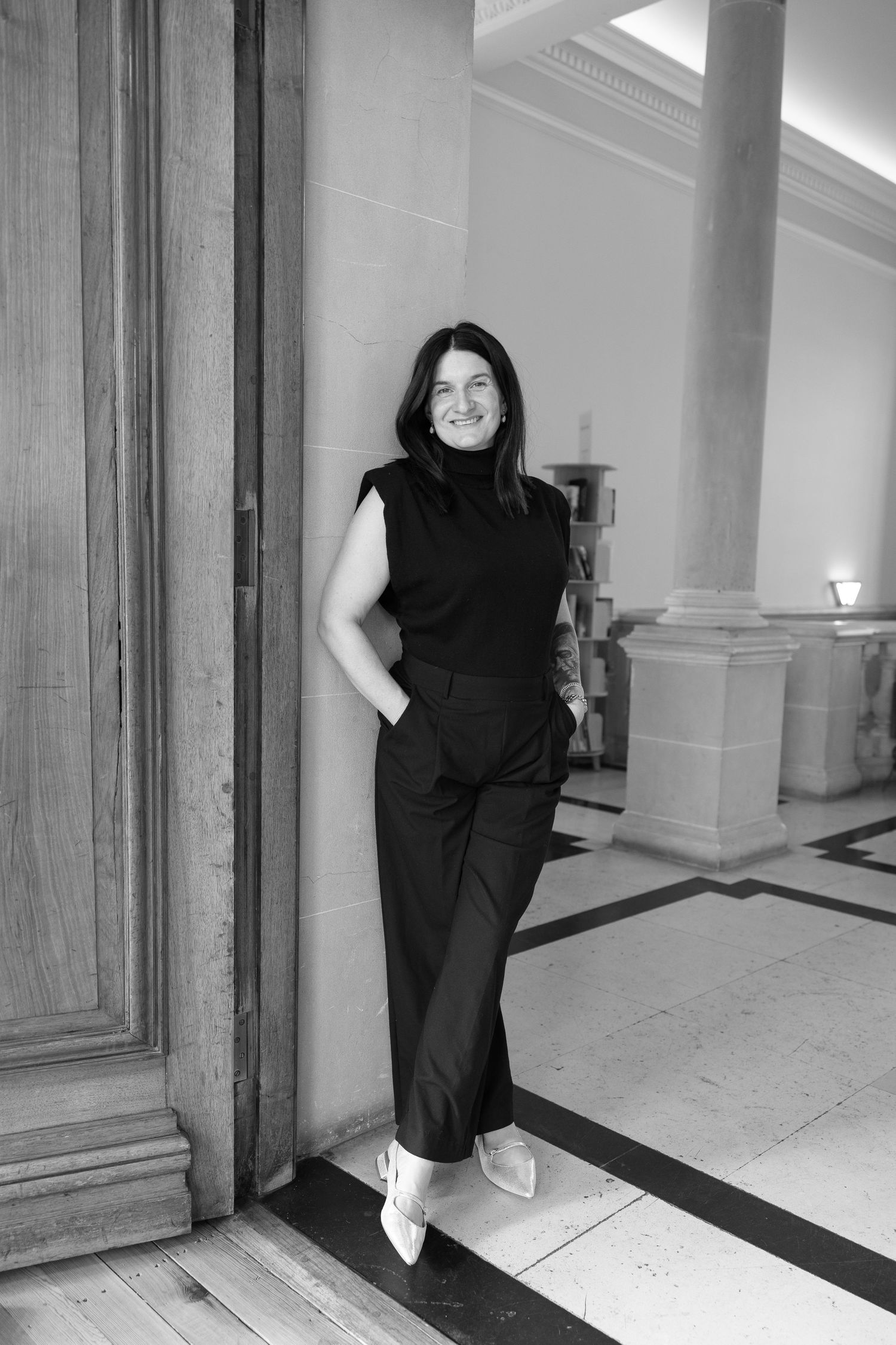Black and white portrait of someone in a sleeveless turtleneck and trousers leaning against an elegant column in a hallway.