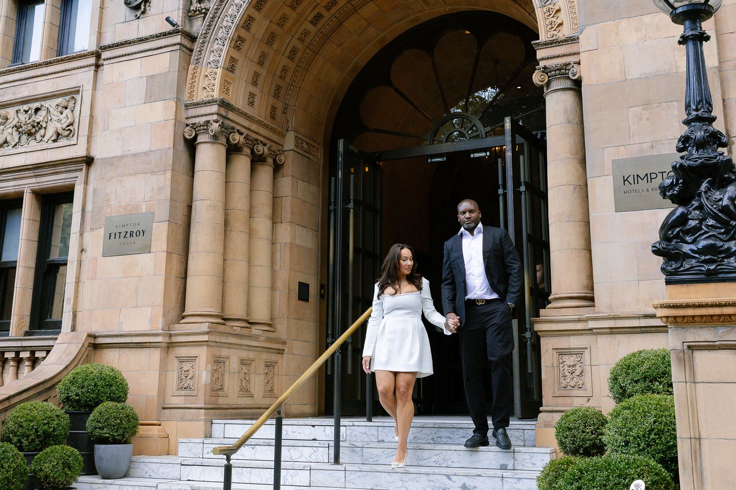 People walk down grand stone steps outside an ornate historic building with manicured topiary bushes.