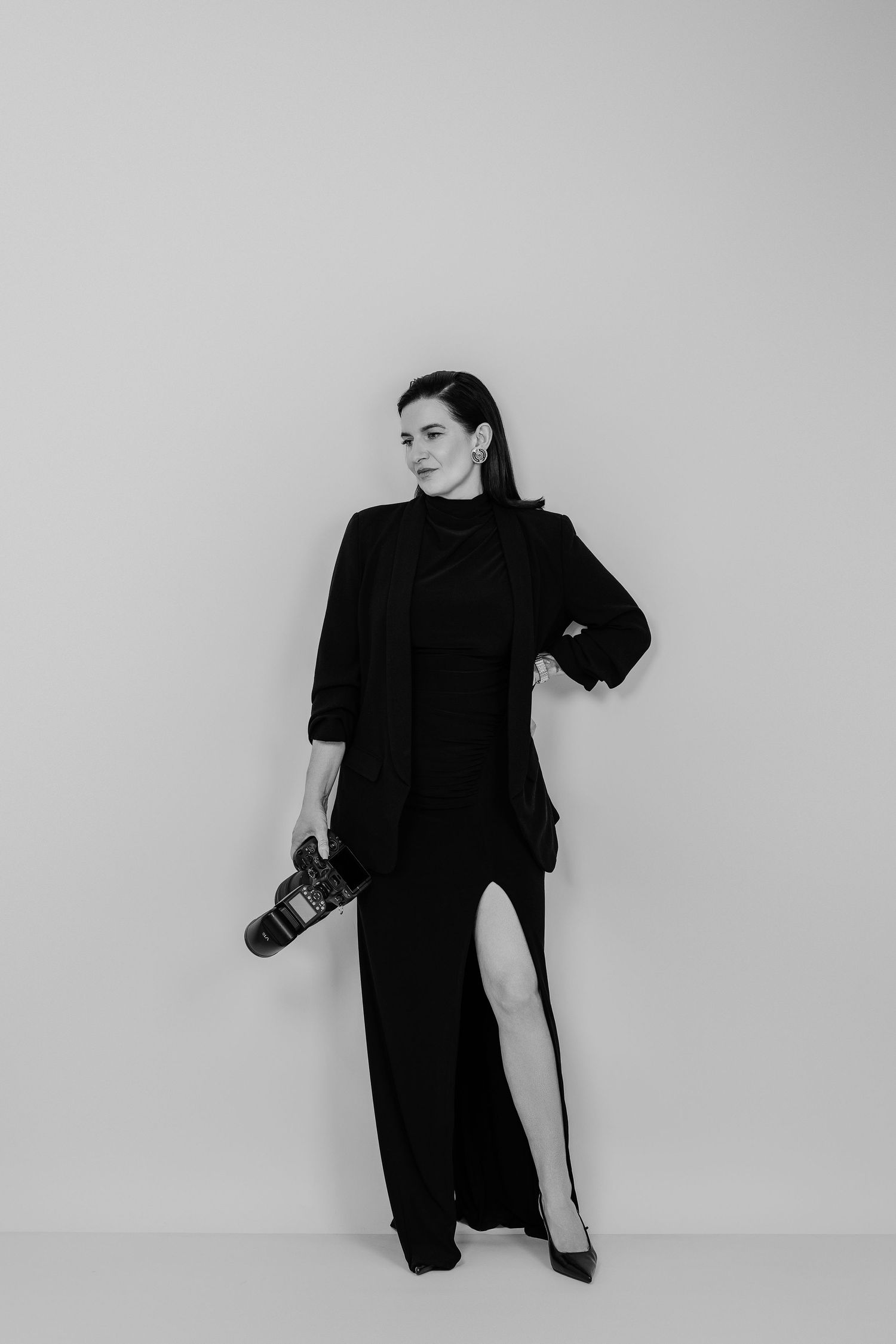 Black and white photo of a woman in elegant black outfit holding a camera, posing confidently against a light background.