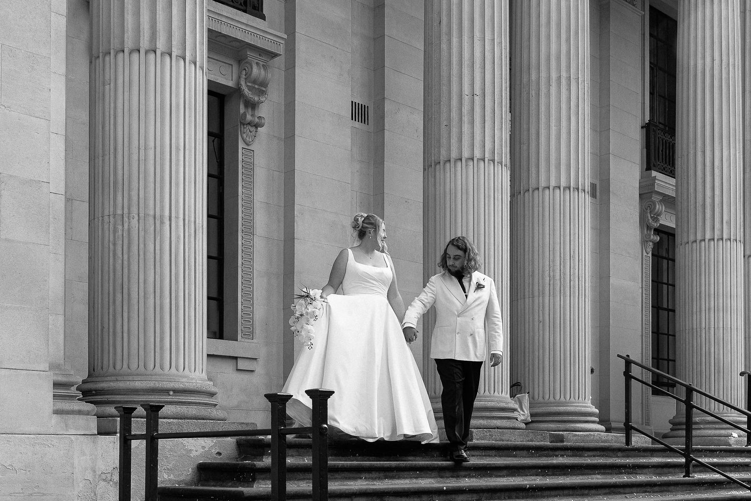 A couple stands on the steps of a grand columned building in black and white wedding attire.