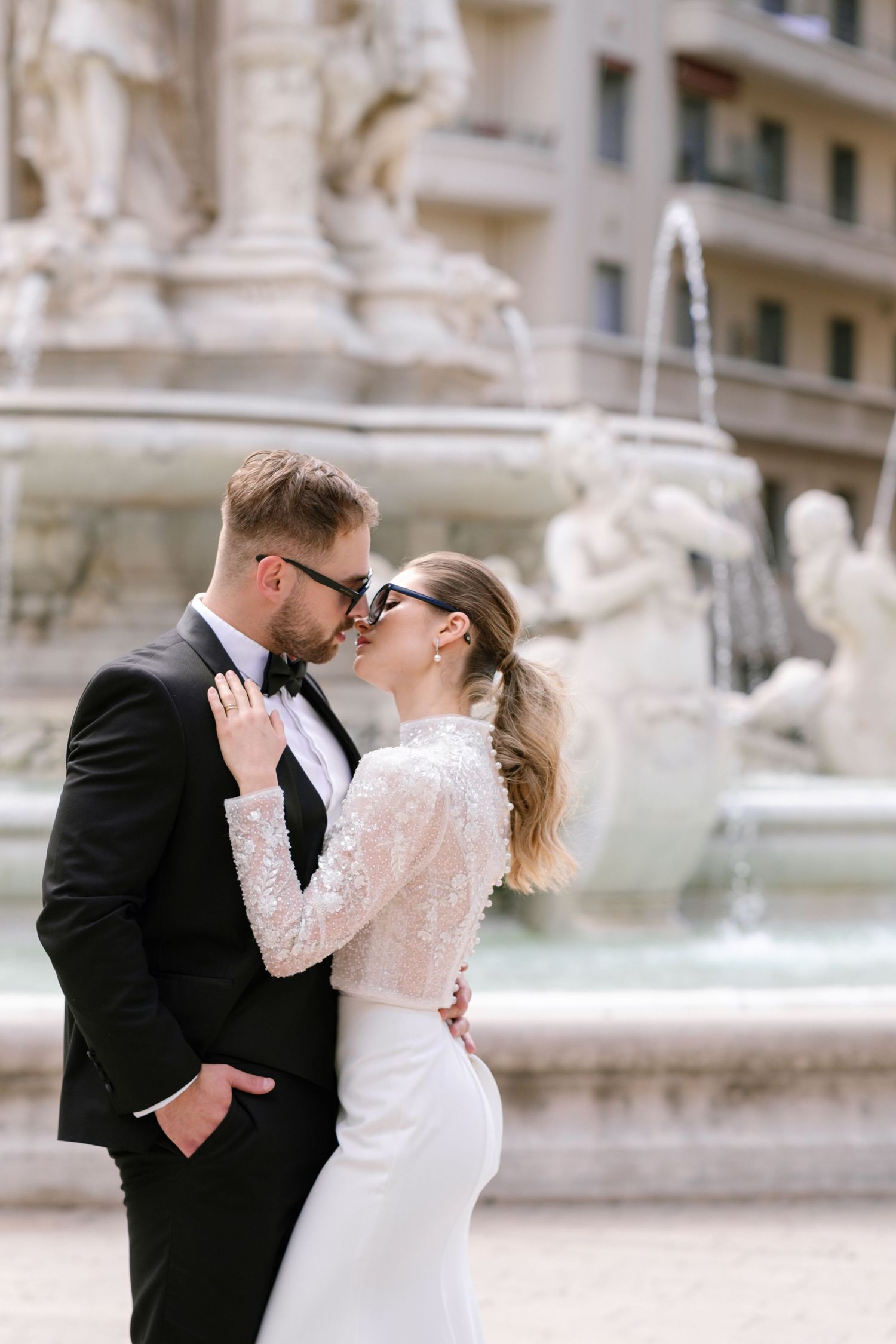 Romantic couple embracing by ornate fountain in formal wedding attire.