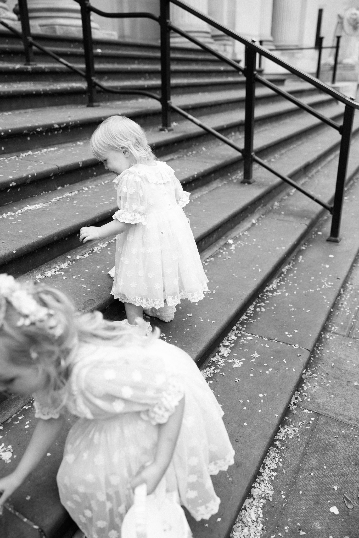 Black and white photo of flower girls in white dresses climbing steps with scattered petals.