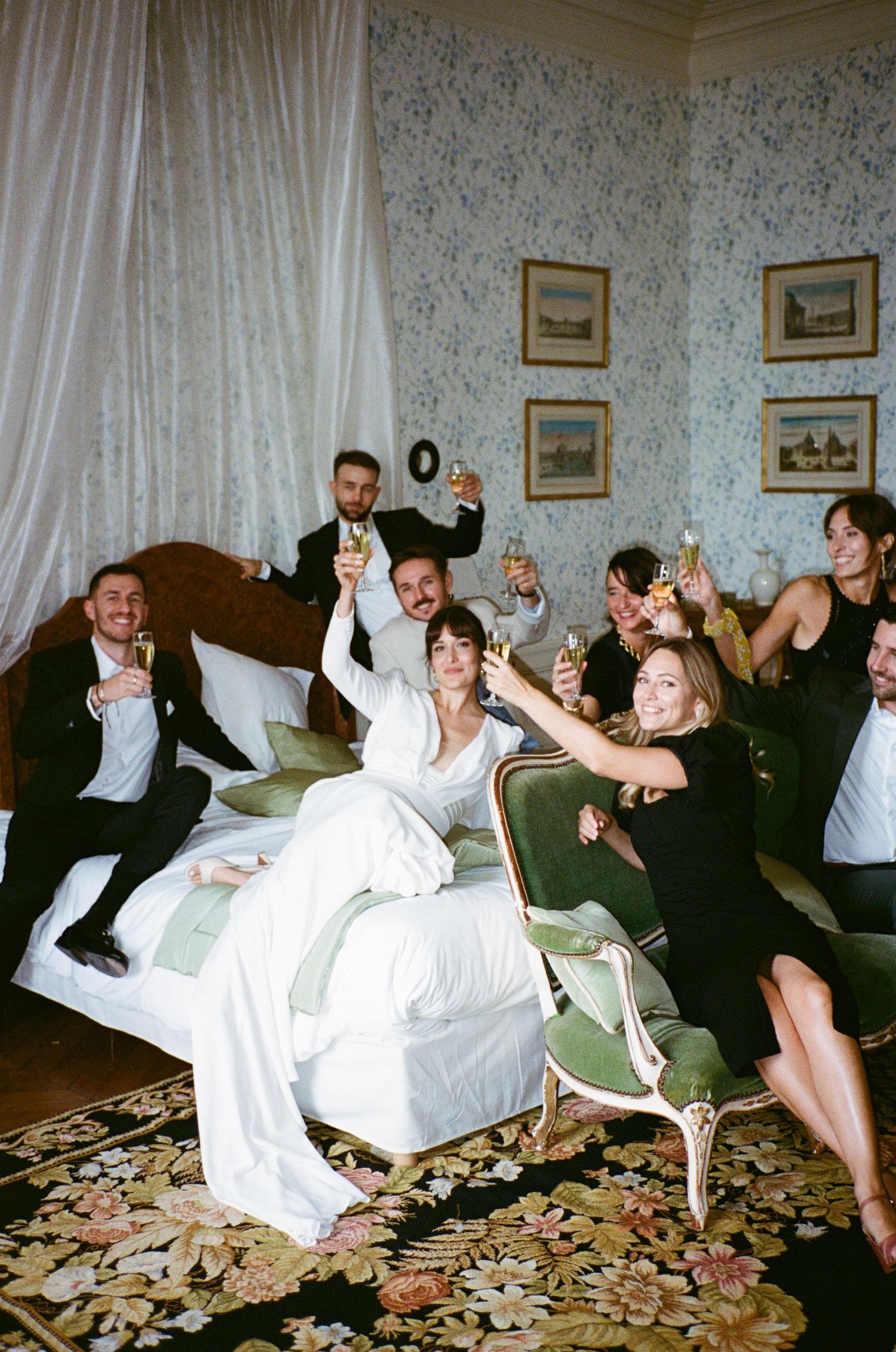 A celebratory group in formal attire raises champagne glasses while lounging on white furniture in a vintage-style room.