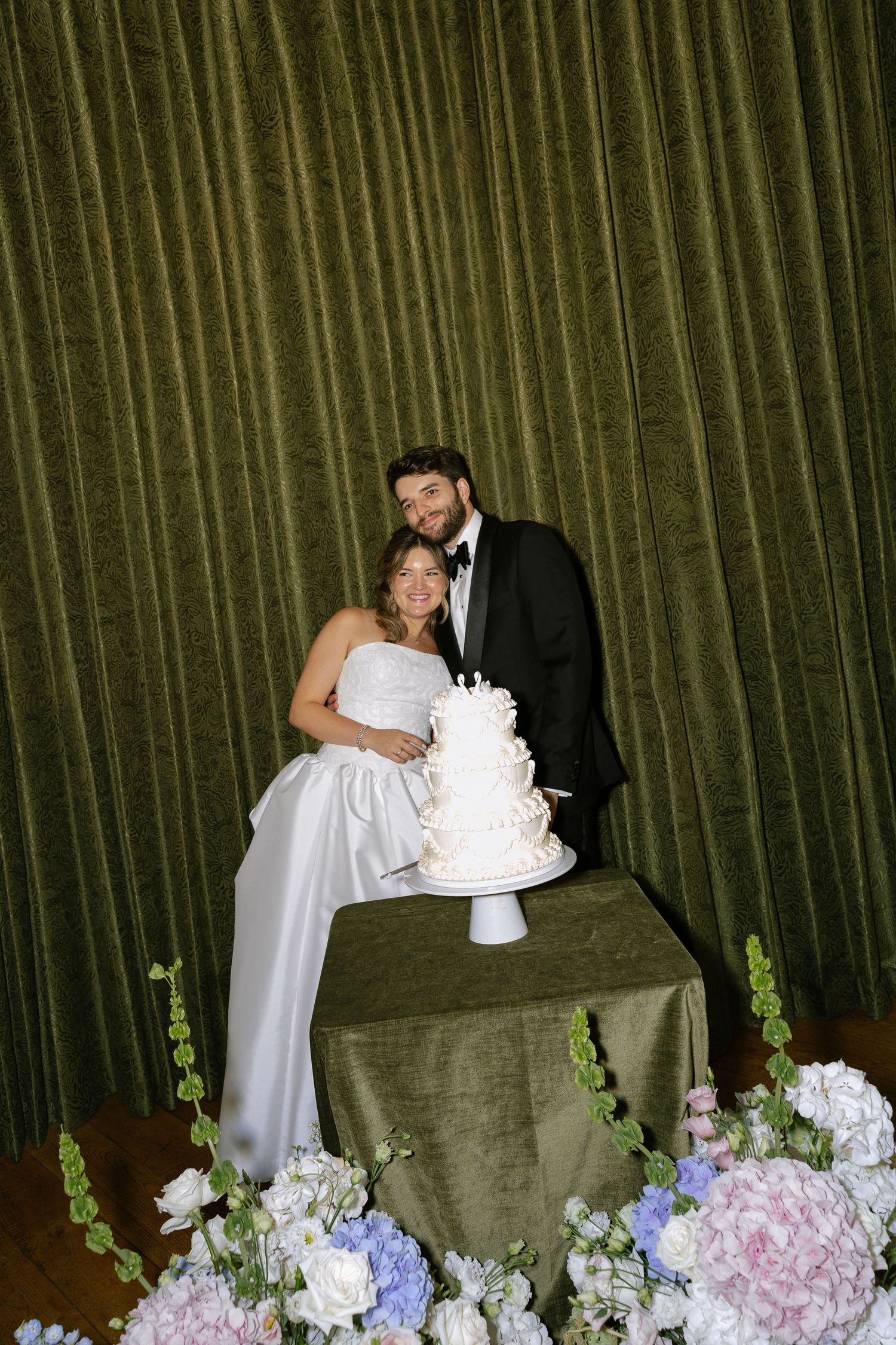 Bride and groom smile beside a white tiered wedding cake on a velvet table with floral arrangements.