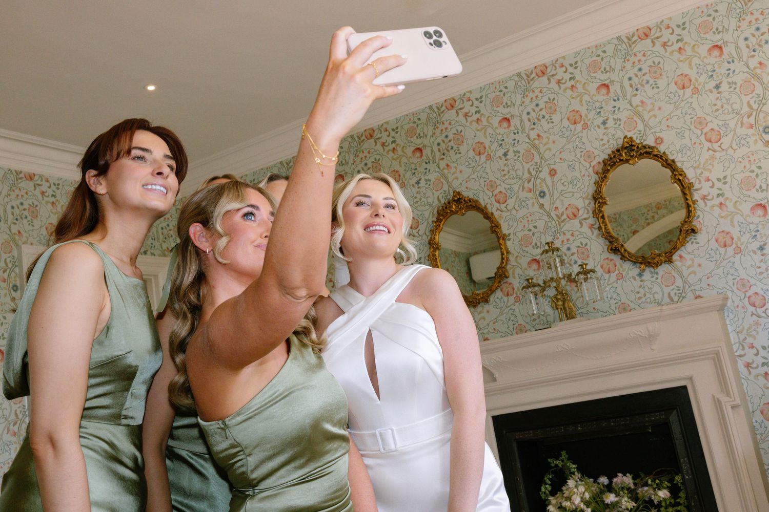 Three attendants in sage green and white dresses take a celebratory selfie in front of floral wallpaper and ornate mirrors.