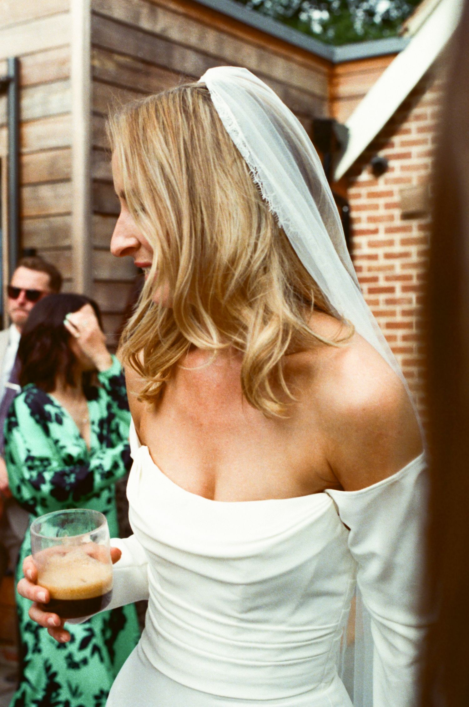 Bride in strapless wedding dress with veil enjoys drink at outdoor reception.