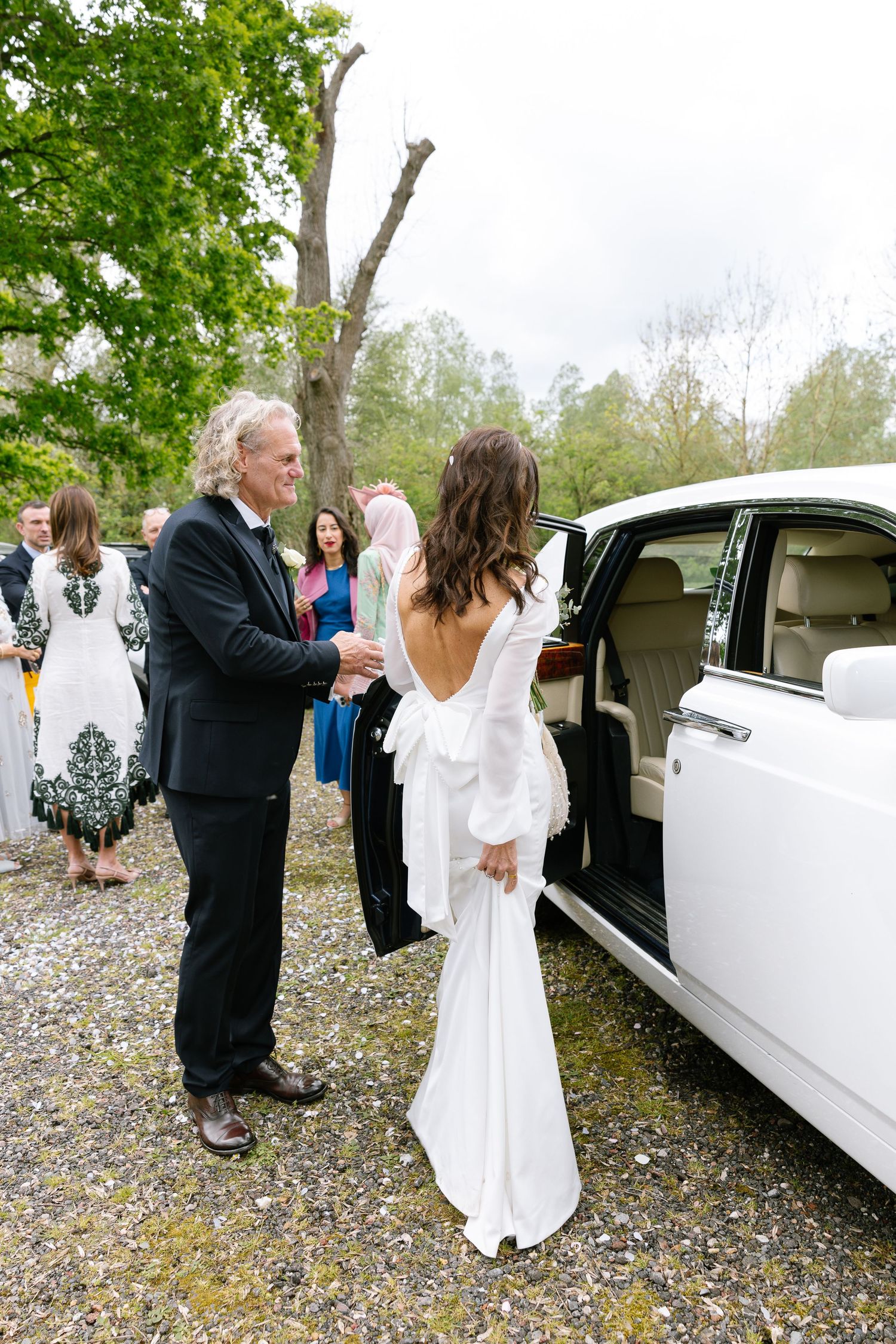 Wedding scene near white limousine with couple and guests on a gravel driveway.