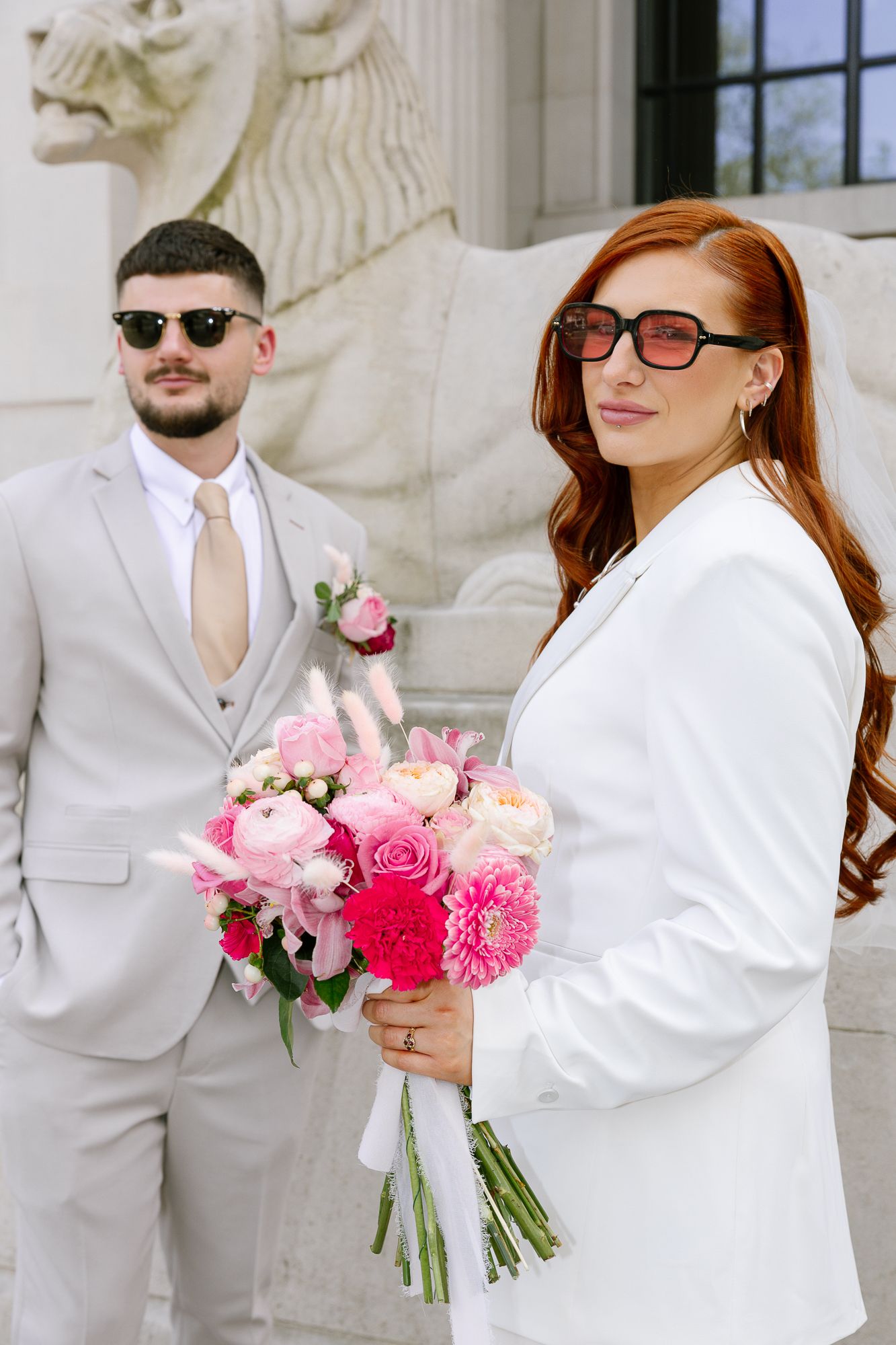 A couple in white and beige attire poses with pink floral bouquet against a stone building backdrop.
