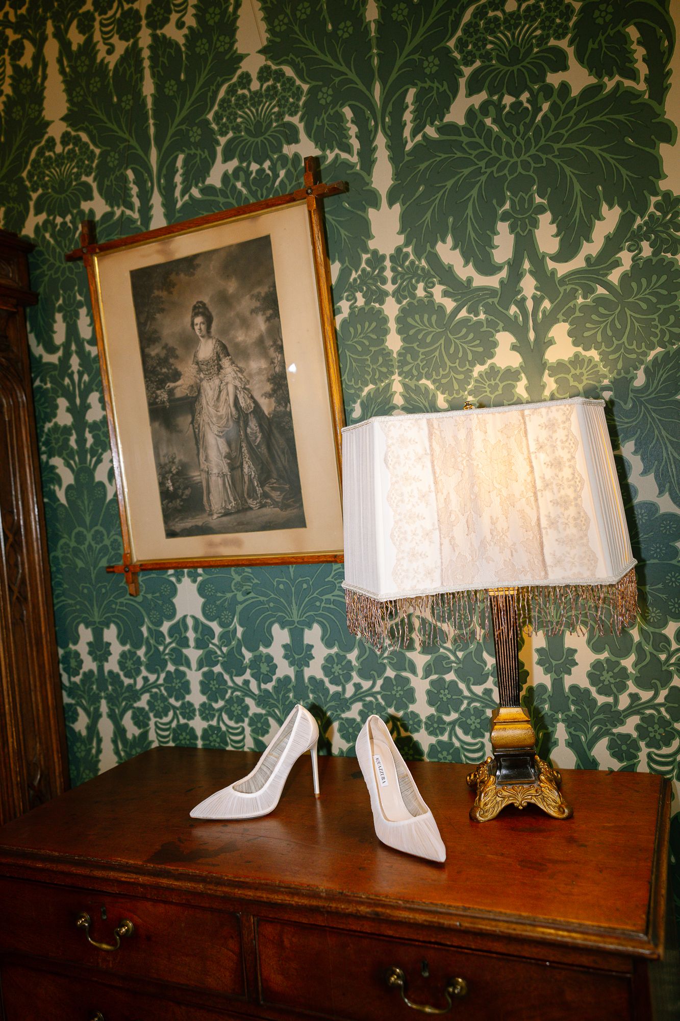 Decorative white lamp shades sit atop an antique wooden console table against green damask wallpaper in an elegant room.